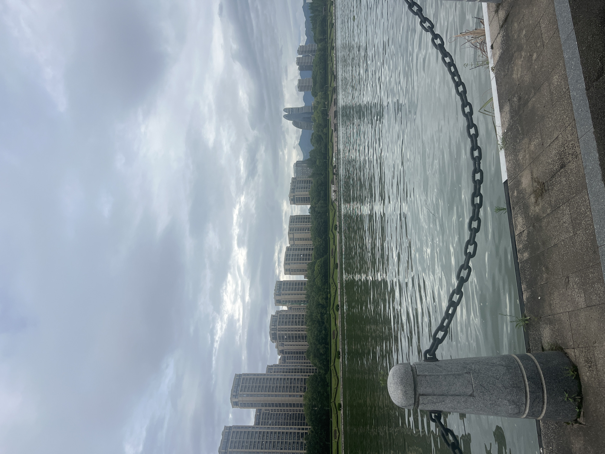 The image shows a waterfront view in Liandu District, China, on a cloudy day. In the foreground, a stone bollard with a thick metal chain attached to it runs along the edge of a paved walkway. Beyond the walkway, a body of water stretches towards the opposite shore, where a line of trees and several high-rise buildings are visible. Further in the distance, more buildings and mountains can be seen under the overcast sky. The water's surface is rippled, reflecting the muted light.