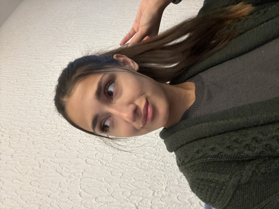 A young adult female is depicted from the upper torso upward, indoors in the city of Zankovići, Montenegro. She has dark brown hair, pulled back into a ponytail, with loose strands framing her face. She is wearing a dark grey top underneath a dark green, open-front cable-knit cardigan. Her gaze is directed towards the upper left of the frame, accompanied by a subtle, closed-lip smile. Her right hand is visible behind her head, positioned near her hair. The background consists of a uniformly lit white wall with a distinct, repetitive, textured pattern resembling stucco or wave-like indentations.