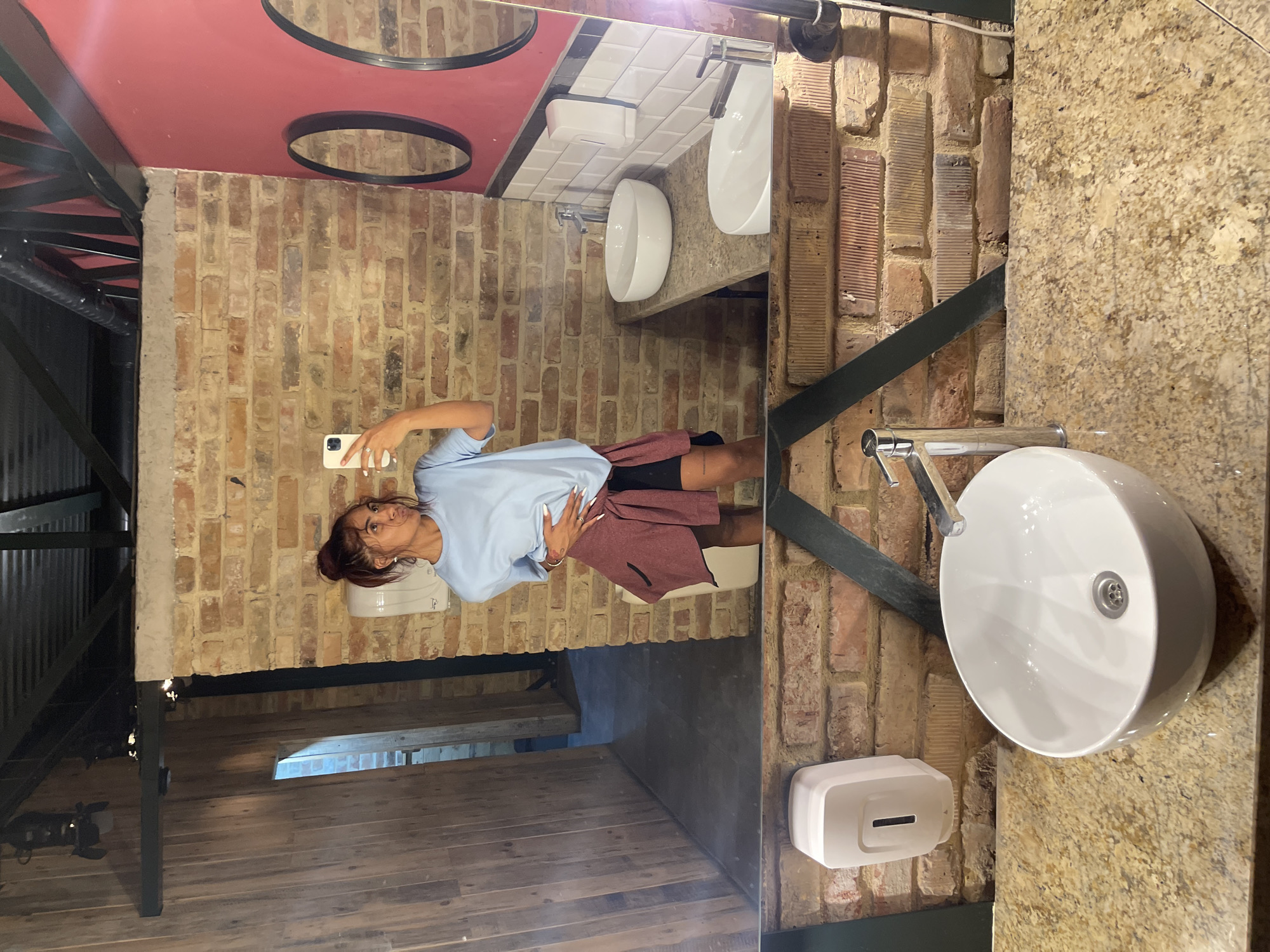 A young woman with auburn hair pulled into a bun takes a mirror selfie in a public restroom. She is wearing a light blue t-shirt and black shorts, with a dark pink cardigan tied around her waist. Her left hand rests on her hip, and she holds a white smartphone in her right hand, capturing the reflection. The restroom has exposed brick walls and a dark, industrial-style ceiling with exposed pipes. In the reflection, two white sinks with chrome faucets are visible on a granite countertop. A white soap dispenser is mounted on the brick wall to the left of the sinks. The overall aesthetic suggests a trendy, urban establishment, possibly a bar or restaurant. The location is Varna, Bulgaria.