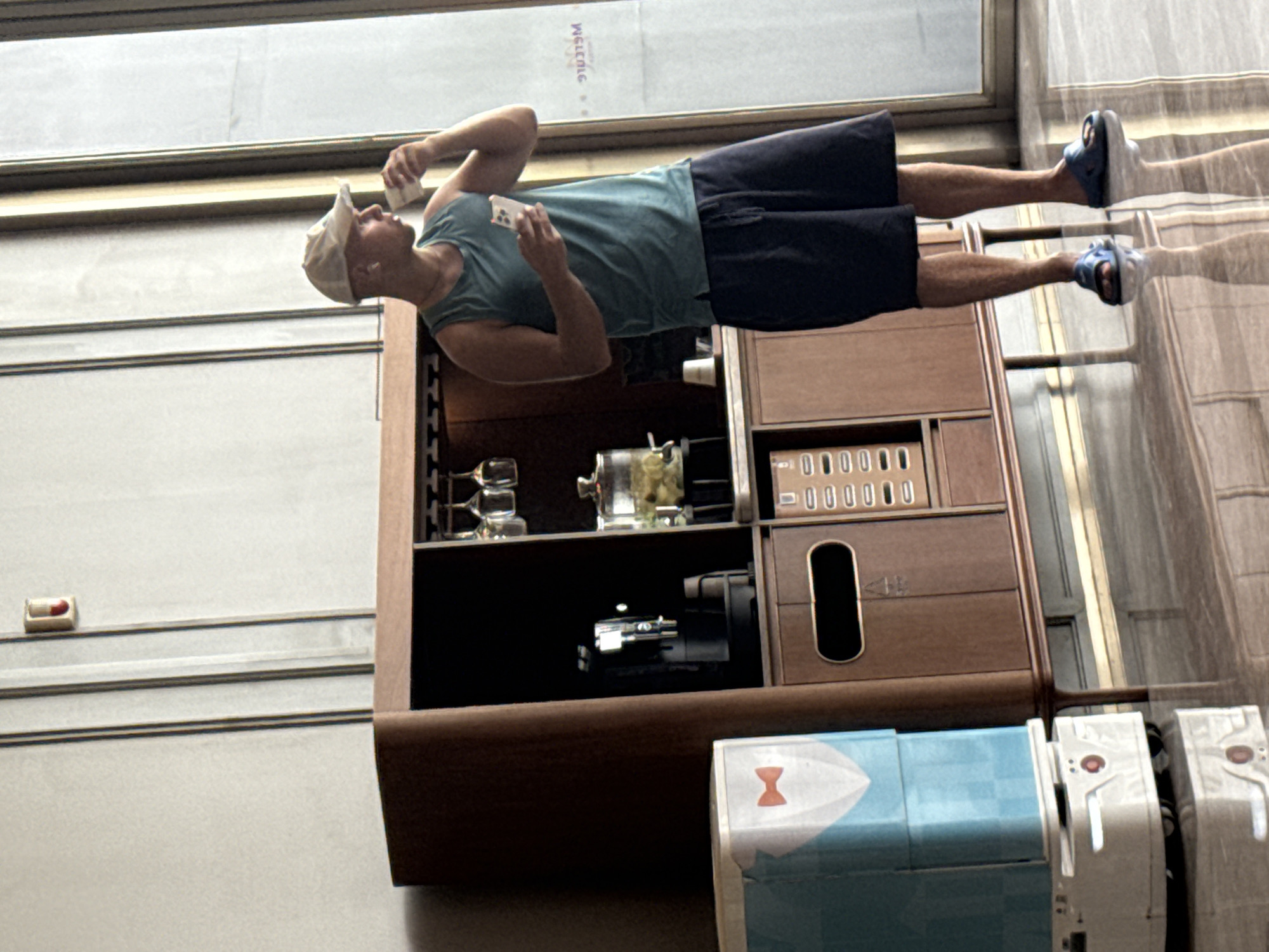 In the Huangpu District of China, a young man wearing a teal tank top, black shorts, and a white baseball cap stands to the right of a modern wooden cabinet. He holds a white cup to his lips with his right hand and a smartphone in his left. The cabinet features a black coffee machine, a glass dispenser filled with yellow items, and wine glass holders. To the left of the cabinet is a light blue and white cylindrical service robot with a red bow tie graphic. The scene takes place indoors with a tiled floor that reflects the objects above.