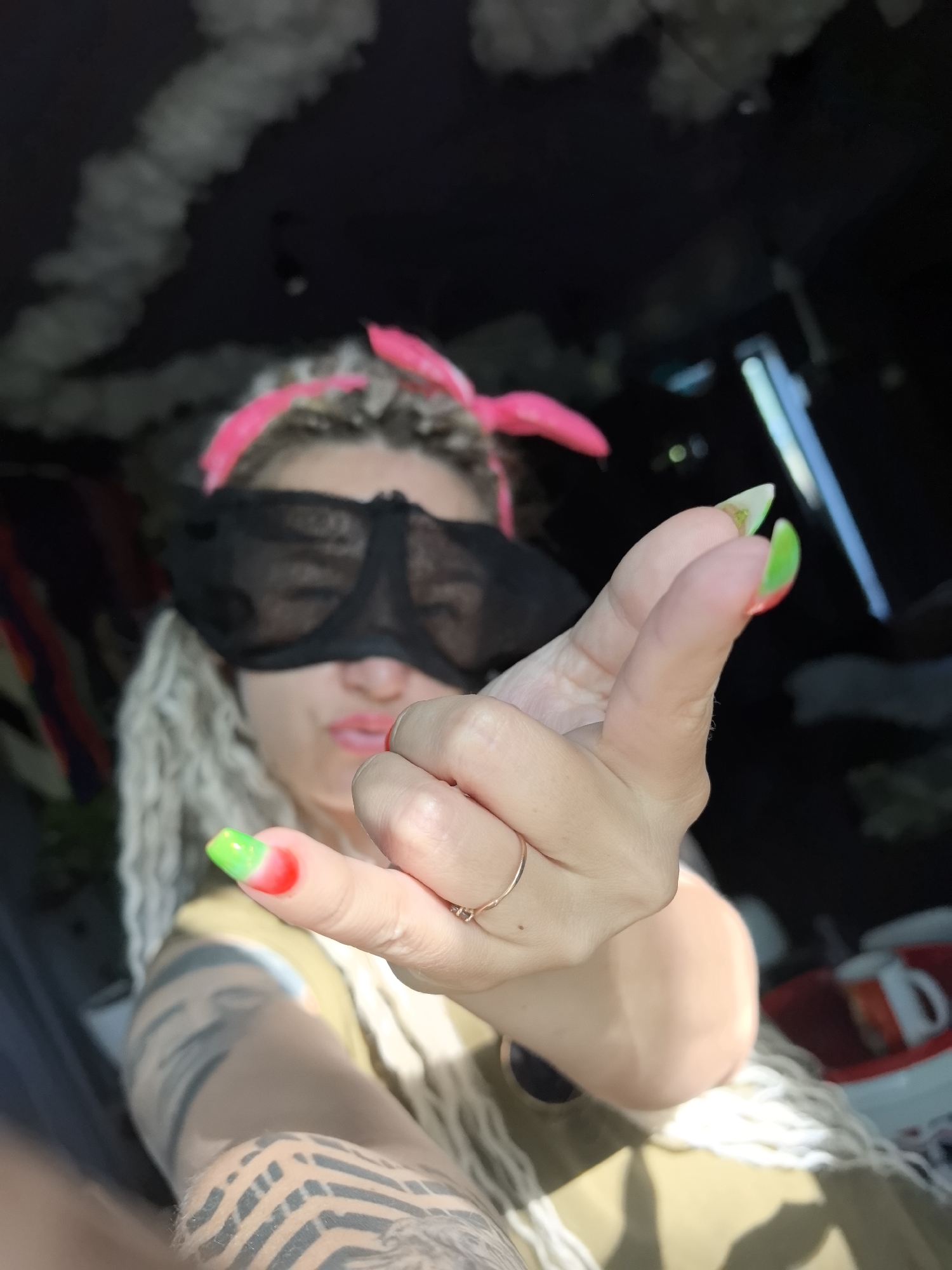 A person with fair skin, long light-colored braided hair, a pink headband, and a black sleep mask covering their eyes is holding up their left hand with the middle finger extended. The nails on the hand are painted with a red and green ombre effect. The person is wearing a ring on their ring finger. They have tattoos on their left arm. The background is out of focus, but it appears to be a window or doorway. There is a red container with a white mug in the background, and a dark ceiling with white decorations above. The location is unknown and there is no basis to assume it is Noviy, Russia.