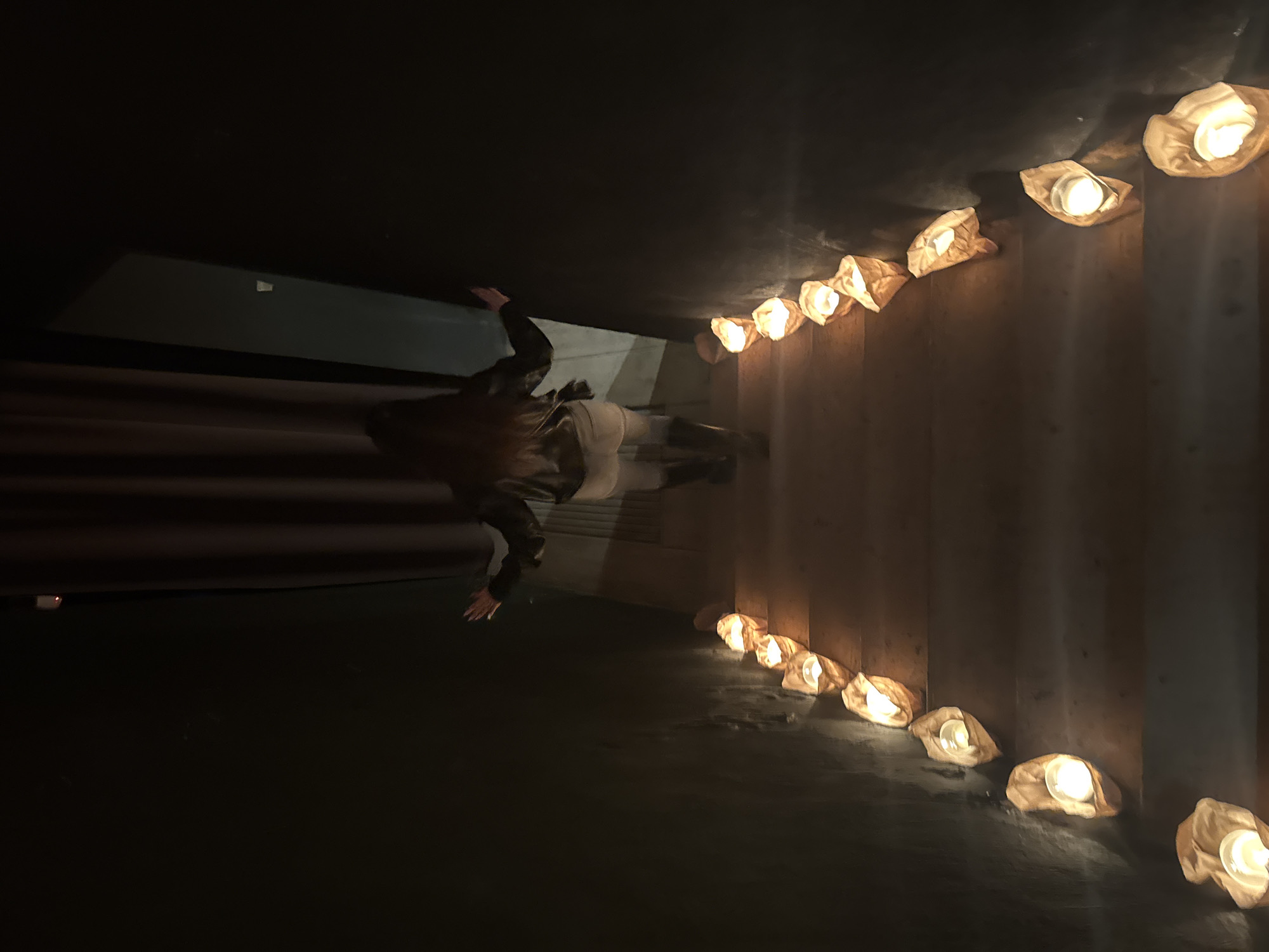 The image, originally rotated 90 degrees counter-clockwise, depicts a person moving through a dark, enclosed architectural space illuminated by a series of luminaries.
In the mid-ground, a lone individual is visible, their figure blurred, suggesting motion. They appear to be wearing a dark top or jacket and lighter-colored trousers or a skirt, with long, dark hair. One arm is raised, possibly touching a wall or railing as they proceed.
Along the right side of the frame, a continuous line of approximately a dozen glowing luminaries is arranged. These appear to be white paper bags, each containing a light source, likely a candle or LED, emitting a warm, soft yellow-orange glow. The lights cast reflections and highlights on the adjacent surfaces.
The setting is characterized by dark, solid walls, possibly concrete or a similar material, which create a tunnel-like or underpass effect. Horizontal bands or architectural features are faintly discernible behind the person. The floor or a low-lying surface in the foreground is dark and somewhat reflective.
The scene takes place at night or in a deeply shaded interior, as indicated by the reliance on artificial lighting from the luminaries. The warm glow of the lights creates an inviting, yet somewhat mysterious, ambiance within the otherwise dark environment. There is no visible text in the image. This scene is located in Sofia, Bulgaria.