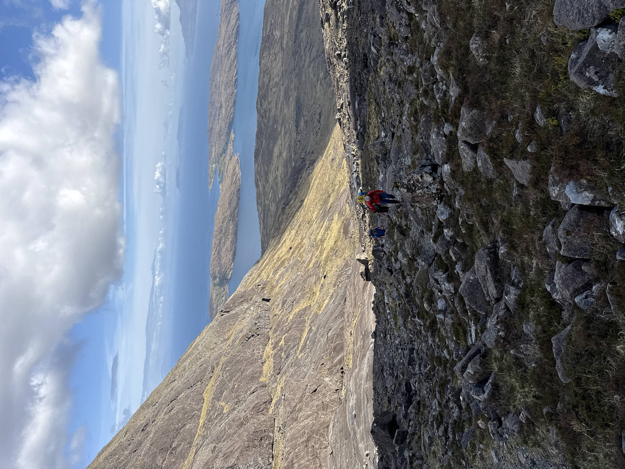 The image shows a mountain landscape. Two figures, one wearing a red jacket and a backpack, are situated on a steep, rocky slope. The terrain is rugged with patches of grass. In the background, there is a view of water, islands, and a cloudy sky. The location is Unknown, Unknown.