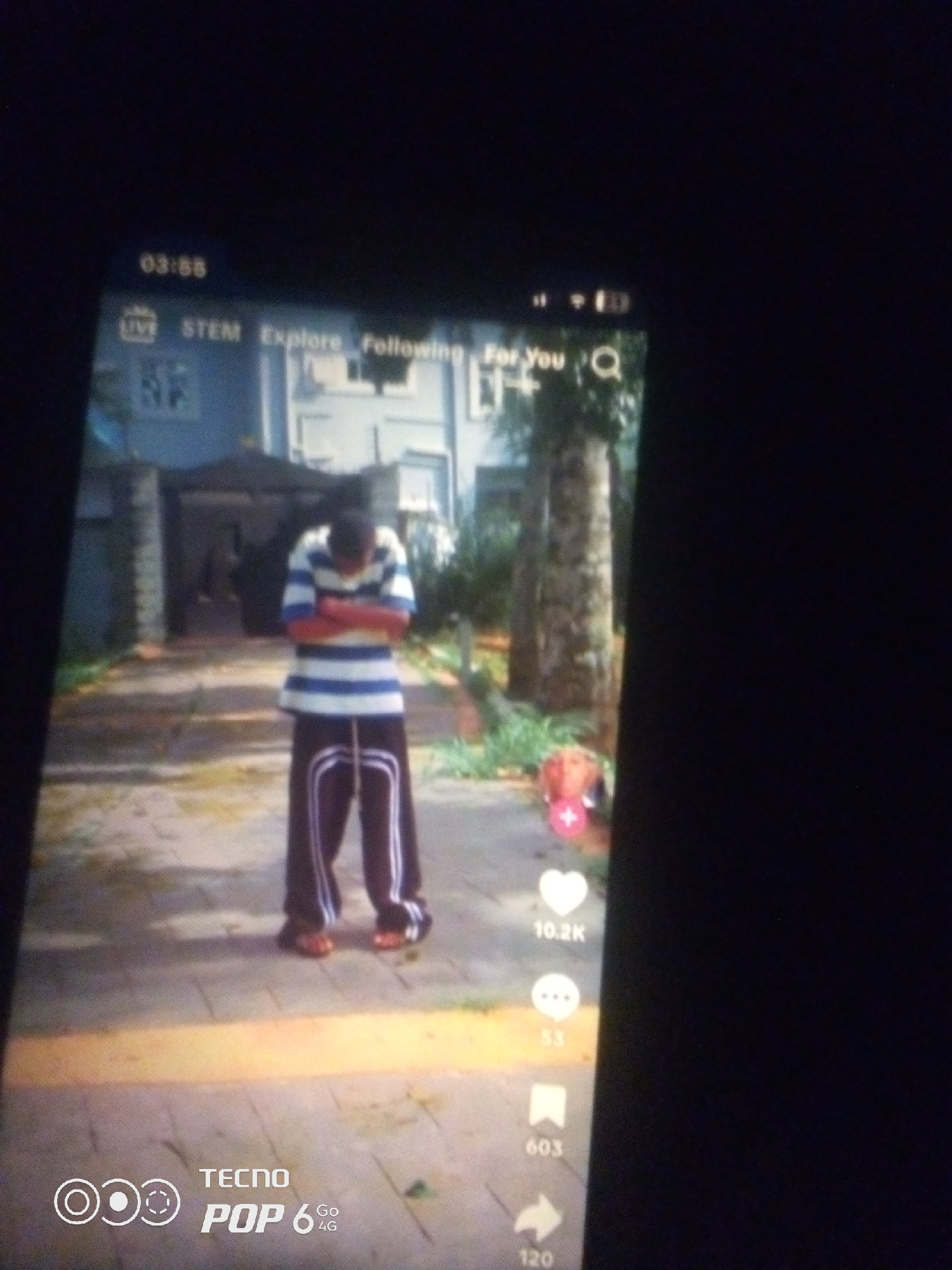 The image displays a smartphone screen with a video playing. The video shows a young person standing outdoors in what appears to be a residential area. The person is wearing a horizontally striped shirt and dark pants with white stripes. They are facing away from the camera with their arms crossed. Behind them, a building with a light-colored facade and a driveway can be seen. The screen also shows social media interface elements, including icons for live, STEM, explore, following, and a search magnifying glass, as well as engagement metrics like "10.2K" likes and "53" comments. The bottom of the screen shows "TECNO POP 6 4G". The setting appears to be a daytime scene. There is no visual information in the image that allows for the specific location of Howayi, Nigeria to be identified.