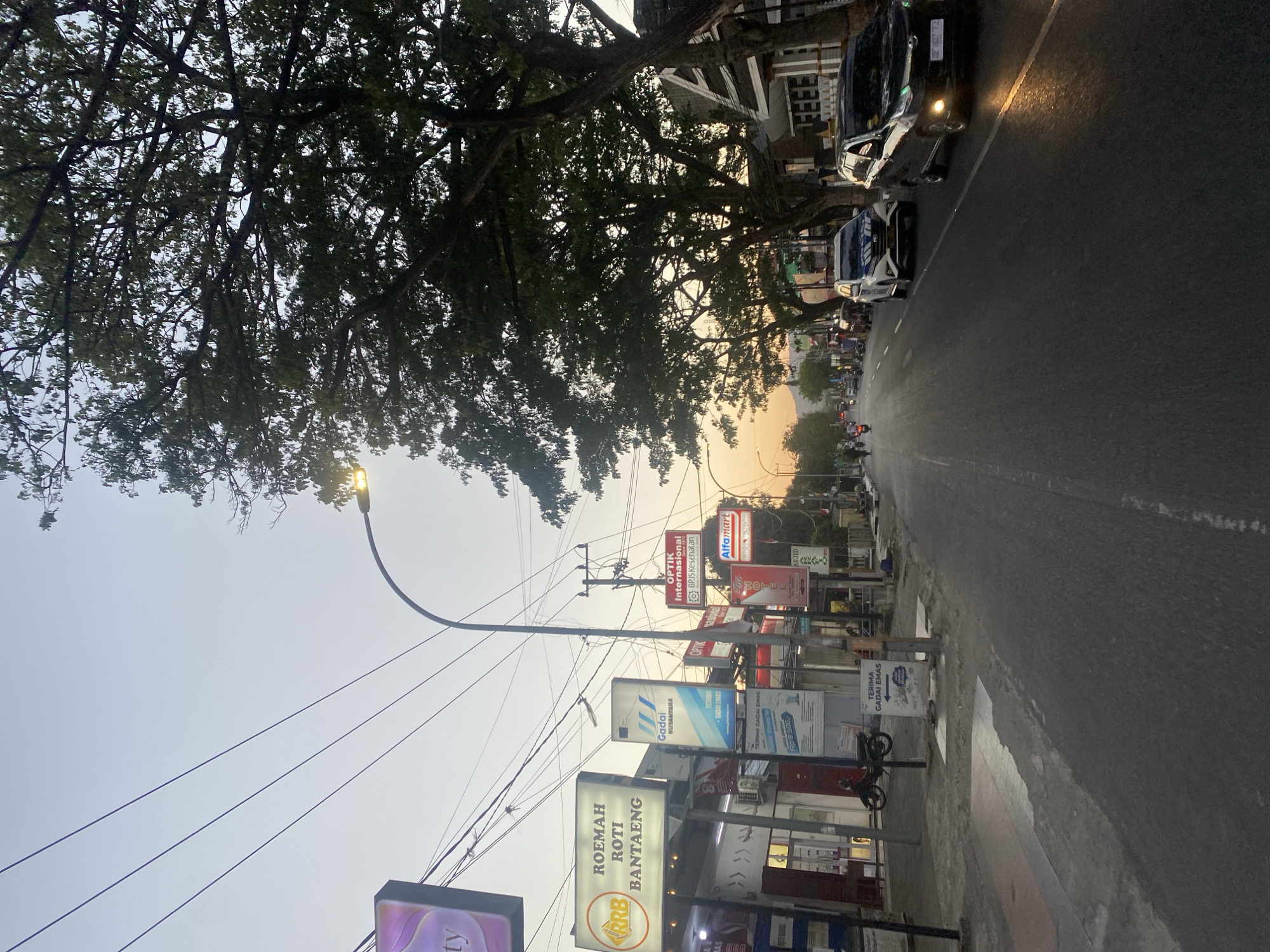 The image depicts a street scene in Bantaeng, Indonesia, during twilight. The sky is a pale blue, transitioning to a warm orange glow near the horizon, suggesting either sunrise or sunset. Large trees with dense foliage line the right side of the street, casting shadows. Several businesses are visible along the left side of the road, indicated by illuminated signs. Notable signs include "ROEMAH ROTI BANTAENG" (Bantaeng Bread House), "Optik Internasional" (International Optician), and "Alfamart," a convenience store chain. There are utility poles with numerous overhead wires crisscrossing the sky. Vehicles, including cars and motorcycles, are present on the asphalt road, which has painted white lines. Some people are visible as distant figures on the street. The overall environment appears to be a commercial street with residential buildings in the background.