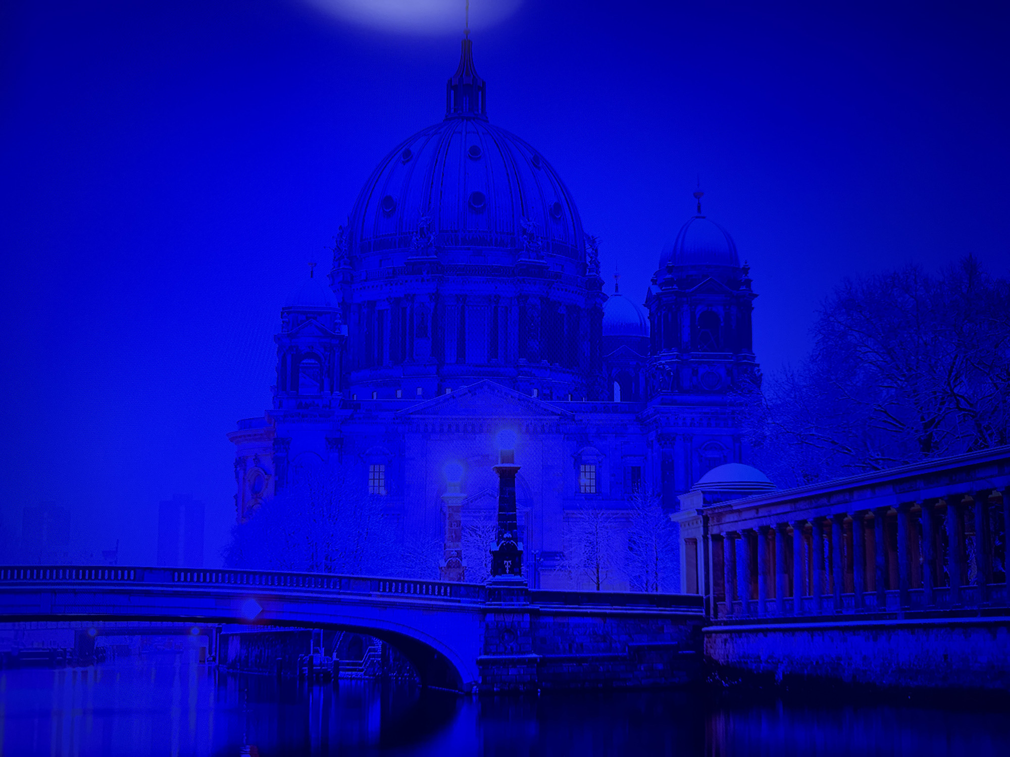 The image depicts a nighttime view of a large, ornate building with a dome, situated on the bank of a body of water. A bridge spans the water in the foreground, with lampposts visible along its railing. The building appears to be illuminated, with lights shining from its windows. The surrounding trees are bare and covered in snow, suggesting a winter season. The overall color palette is dominated by shades of blue, creating a cool and atmospheric effect. The image does not depict the city of Pastavy, Belarus.
