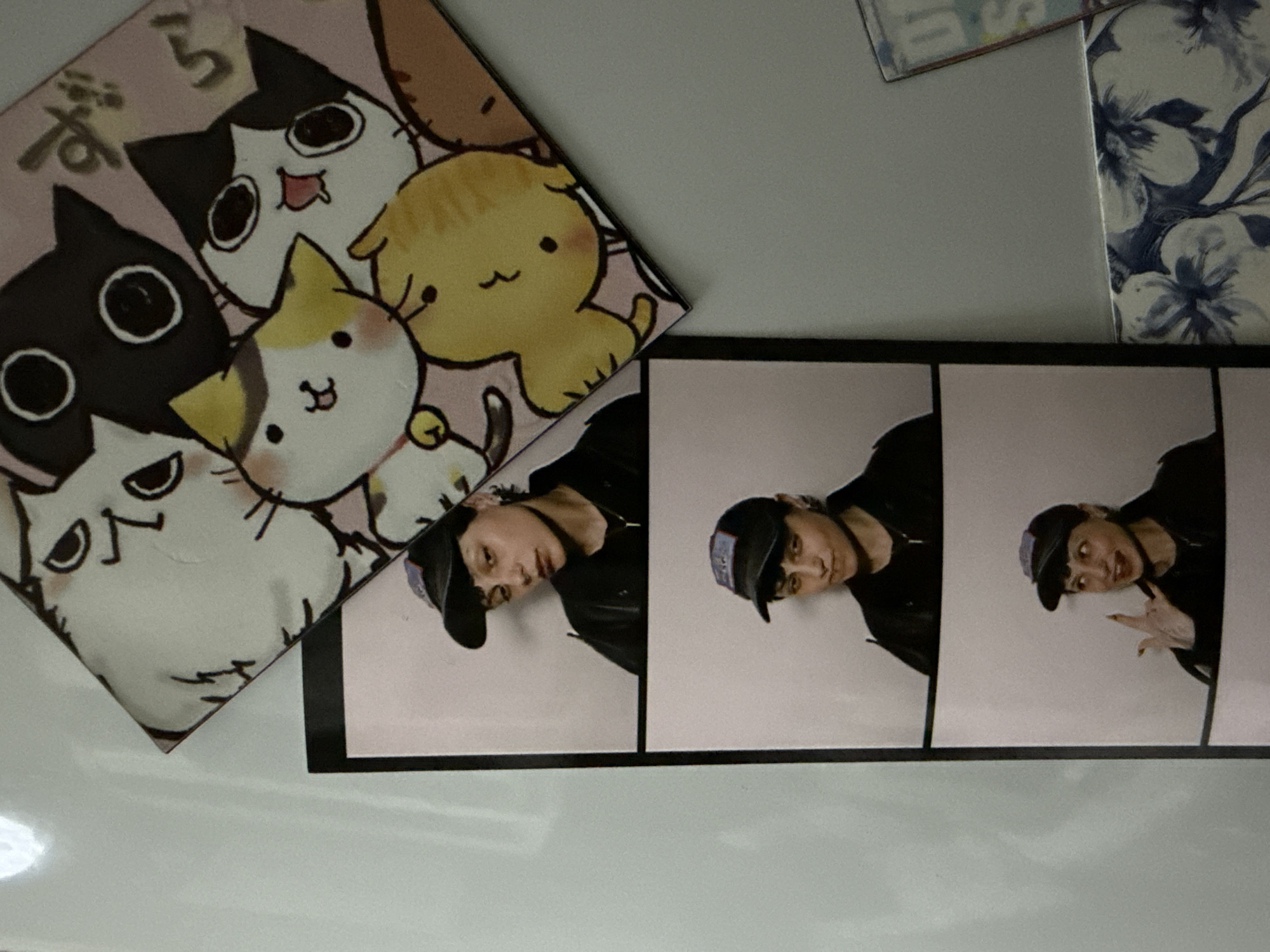 The image shows a close-up of a surface, likely a wall or table, with several items attached or placed on it. On the left, there's a square sticker or print featuring cartoon cats. Adjacent to this, and slightly above, is a strip of three black-and-white photo booth images. Each photo features a person with dark hair wearing a baseball cap and a dark jacket. In the third photo, the person is making a hand gesture. In the upper right corner, there's a partial view of a floral-patterned item or object. The setting cannot be determined to be Saint Petersburg, Russia from the information available in the image.