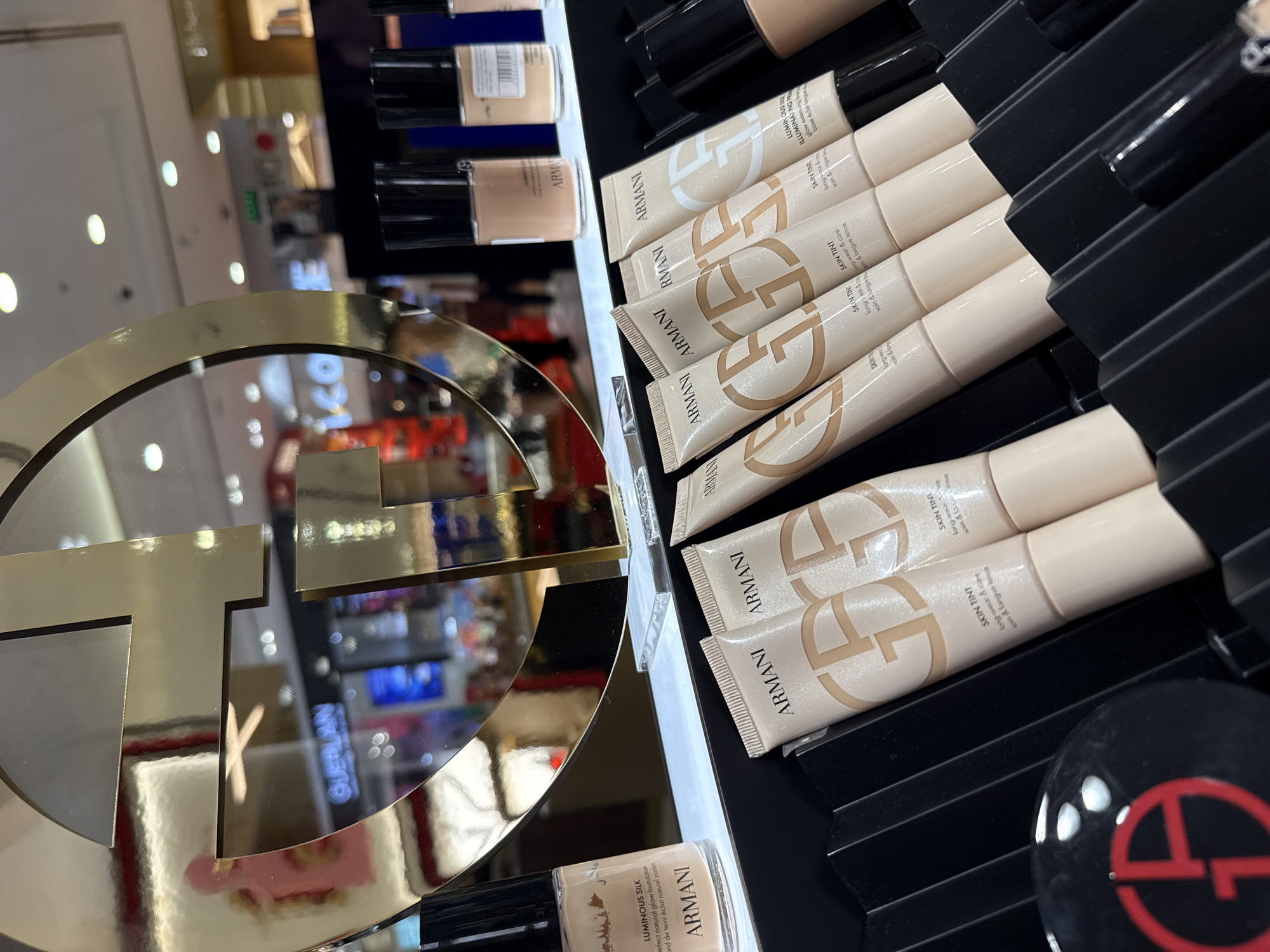 The image displays a close-up view of a retail display featuring Giorgio Armani cosmetic products, specifically foundation and skin tint tubes, arranged in black tiered holders. In the background, a large, golden, stylized 'G' logo is prominent, with reflections of store lighting and merchandise visible on its surface. A bottle of Armani Luminous Silk foundation is positioned on the left side of the display. The overall scene suggests a high-end beauty store or department. The location is specified as Astana, Kazakhstan.