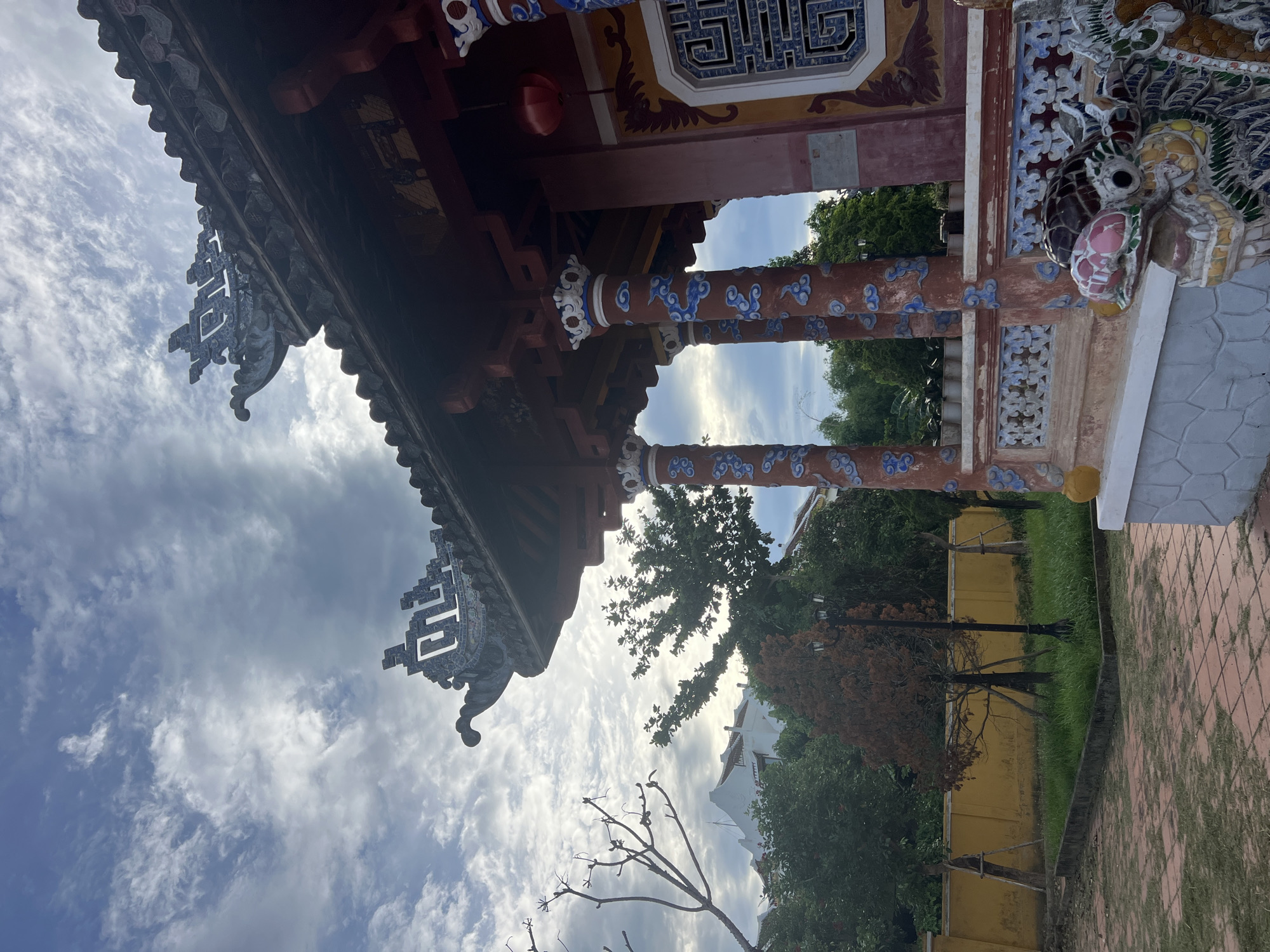 This is a low-angle, medium shot of the exterior of a traditional Vietnamese temple building in Hoi An, Vietnam. The building features a dark, tiled roof with ornate, upturned eaves. Several red columns with blue cloud-like motifs support the roof. The building's exterior walls are painted a faded pink and adorned with decorative window grilles. In the foreground, a colorful ceramic dragon statue is partially visible. The courtyard in front of the building is paved with red tiles, and to the left, there's a yellow wall. Lush green trees and a clear blue sky with scattered clouds fill the background.