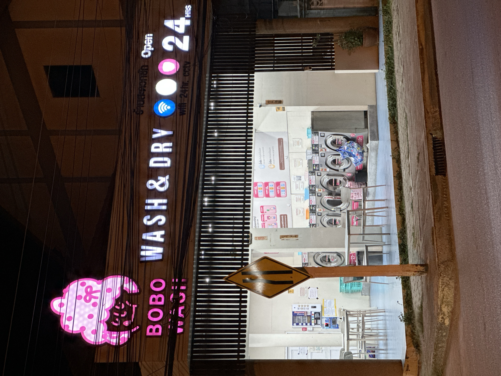 This is a night-time, eye-level, exterior shot of a commercial establishment identified by signage as "BOBO WASH & DRY" located in Baan Mae Nam, Thailand. The facade is illuminated by neon lights and features a prominent pink cartoon-like logo of a character with a bow. The main signage also reads "BOBO WASH & DRY" in illuminated letters, accompanied by icons representing Wi-Fi, a circle, and a pink circle, followed by "24 HRS". Below this, text states "wifi 24hr. cctv". The establishment appears to be a laundromat, with several washing machines visible through an open, covered area. Inside, there are tables and chairs set up. A diamond-shaped road sign with black stripes indicating a narrowing road is positioned in front of the laundromat. The surrounding environment includes a sidewalk and a paved road.