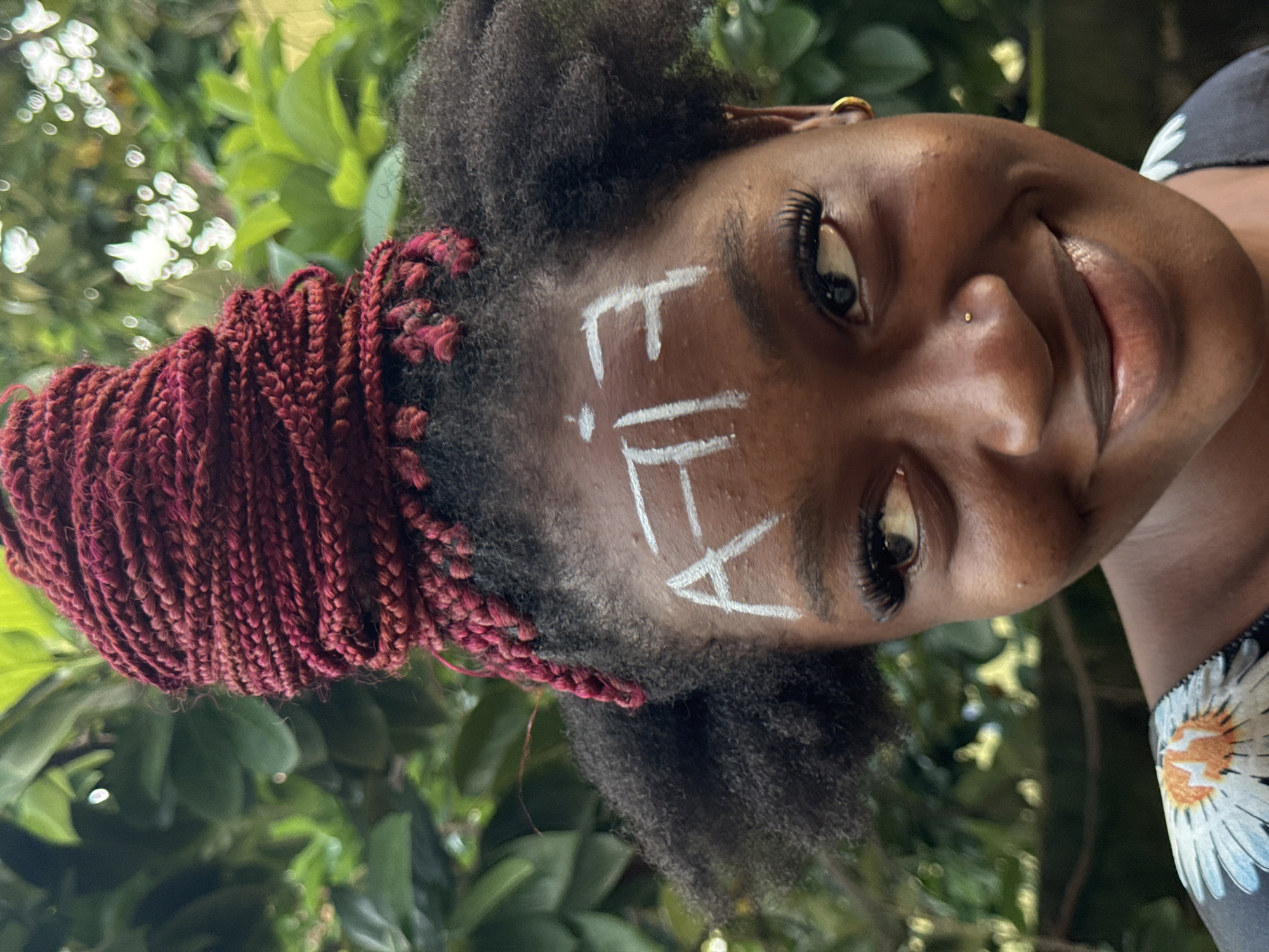 A close-up, head-and-shoulders portrait shows a young Black woman with dark skin, a nose ring, and hoop earrings. She is outdoors, with lush green foliage in the blurred background, suggesting a natural setting possibly within a residential area of Ado, Nigeria. The woman's hair is styled in dark, voluminous afro puffs with a prominent bun of braided dark red hair on top, adorned with dangling braids. Her forehead is painted with white markings that resemble stylized letters or symbols. She is looking off to her right, with a slight smile playing on her lips, and her eyes are accentuated with dramatic false eyelashes. She is wearing a dark garment with a white floral pattern visible on the shoulder. The lighting suggests it is daytime.