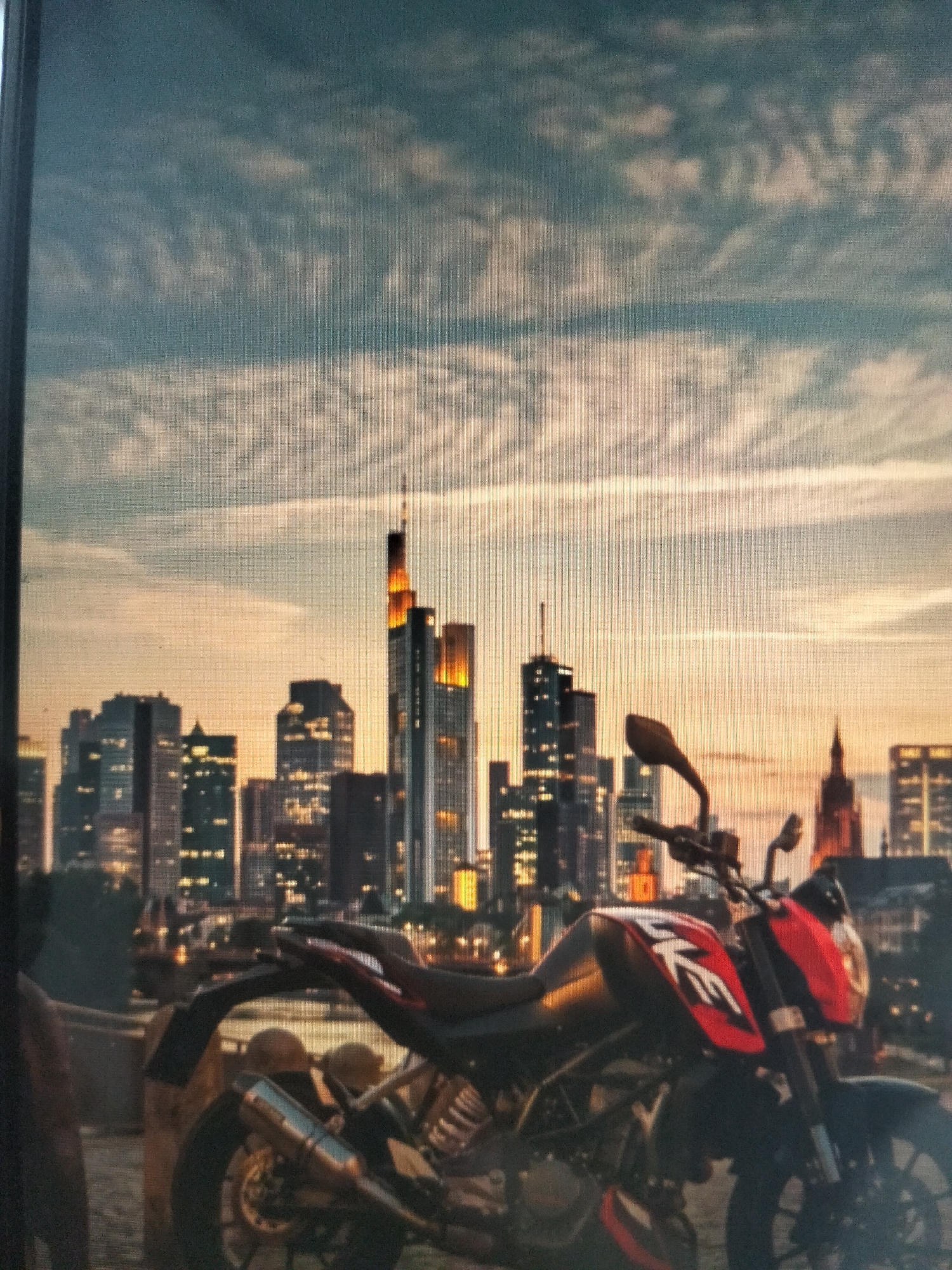 The image displays a red and black motorcycle parked in the foreground, with a city skyline in the background. The skyline features numerous skyscrapers, some of which are illuminated. The sky above is a mix of blue and orange hues, suggesting either sunrise or sunset. The overall scene appears to be a snapshot of an urban environment at dusk or dawn. The location is identified as Payagaboh, Indonesia, though this detail is not visually evident from the image itself. There are no people or specific actions depicted in the scene; it is a still, observational shot.