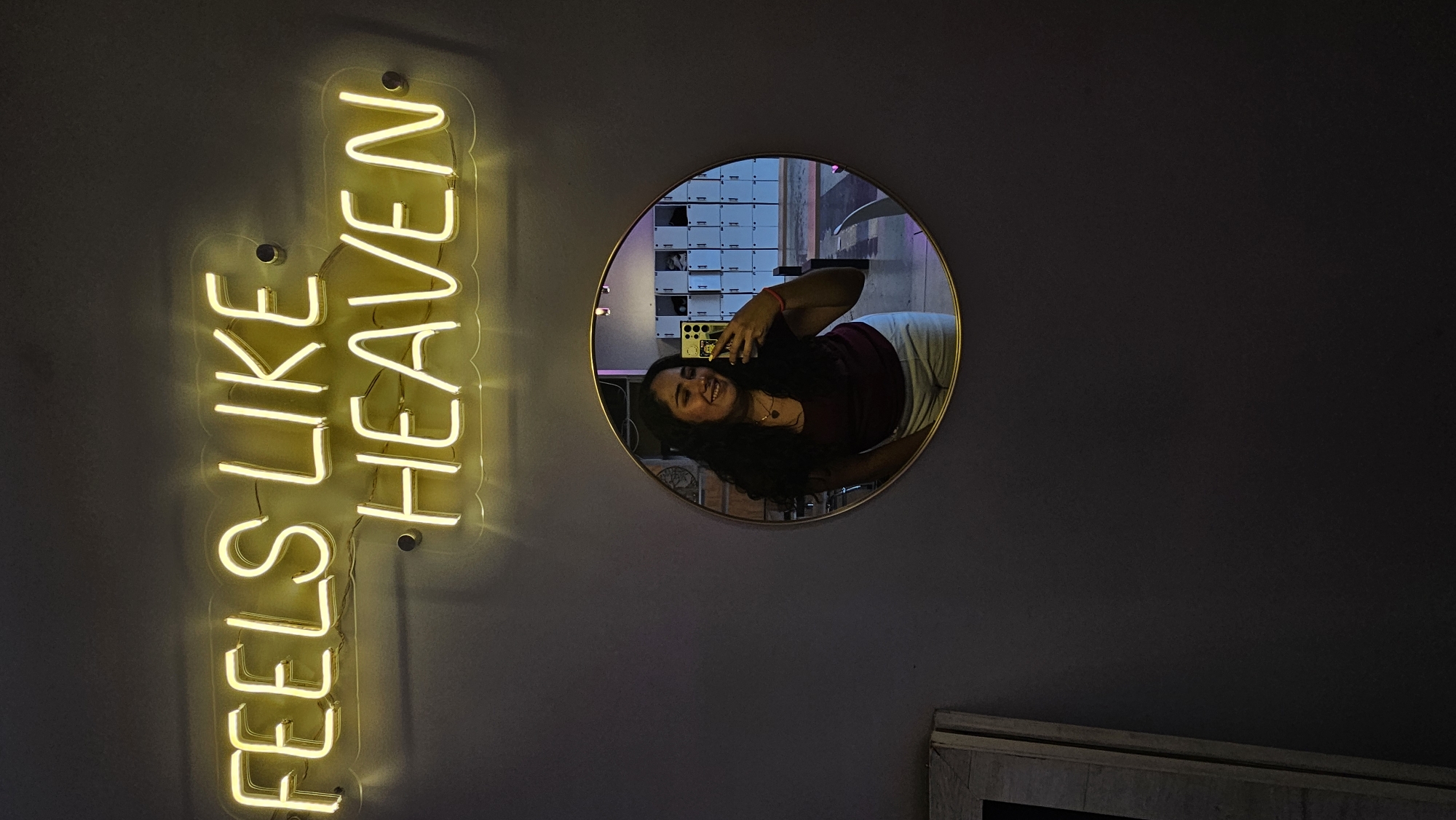 A photograph shows a young woman reflected in a round mirror, positioned on a dark wall. To the left of the mirror is a vertical neon sign that says "FEELS LIKE HEAVEN" in yellow light. The reflection shows the woman holding a phone, presumably taking the picture. Behind her in the mirror, a wall with what appears to be a grid of white compartments is visible. The location is likely in Puebla, Mexico.