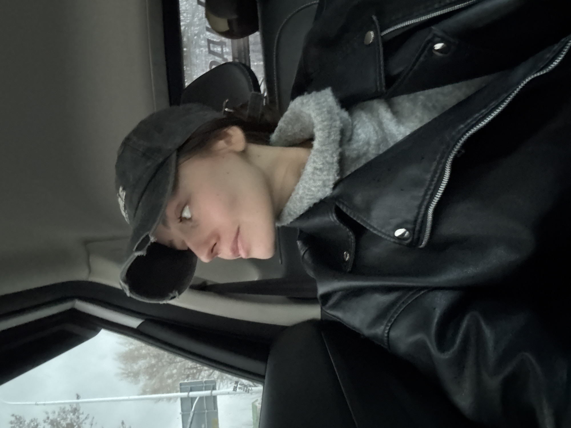 A woman is in the driver's seat of a car. She is wearing a black baseball cap, a grey hooded sweatshirt, and a black leather jacket. The car's window is visible, and the scene outside is a city street. A street sign can be seen. It is not possible to confirm the location is Moscow, Russia from the image.