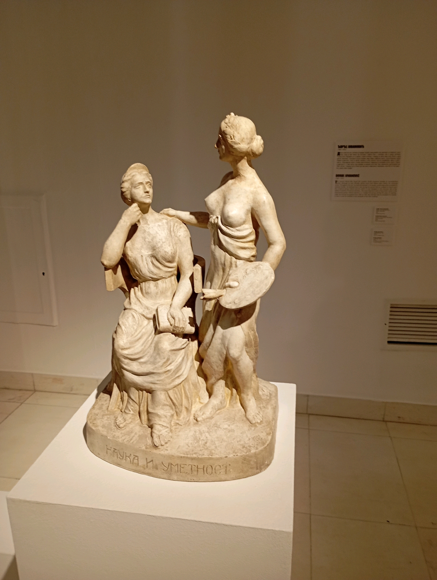 The image shows a marble sculpture displayed on a white pedestal in a museum. The sculpture depicts two female figures. The figure on the left is seated, wearing a draped dress, and holding a book. She is looking upwards with her right hand touching her chin. The figure on the right stands, wearing a toga-like garment and holding a palette and brushes in her left hand. Her right hand is placed on the shoulder of the seated figure. The base of the sculpture is inscribed with text in Cyrillic. The background is a plain, lit wall with a label to the right of the sculpture. The floor is tiled.
Based on the Cyrillic inscription "НАУКА И УМЕТНОСТ" (Science and Art) and the style of the sculpture, this artwork is likely "Science and Art" by Djordje Jovanović, and it is located in the National Museum of Serbia in Belgrade, Serbia. The museum is situated in the Republic Square in Belgrade.
