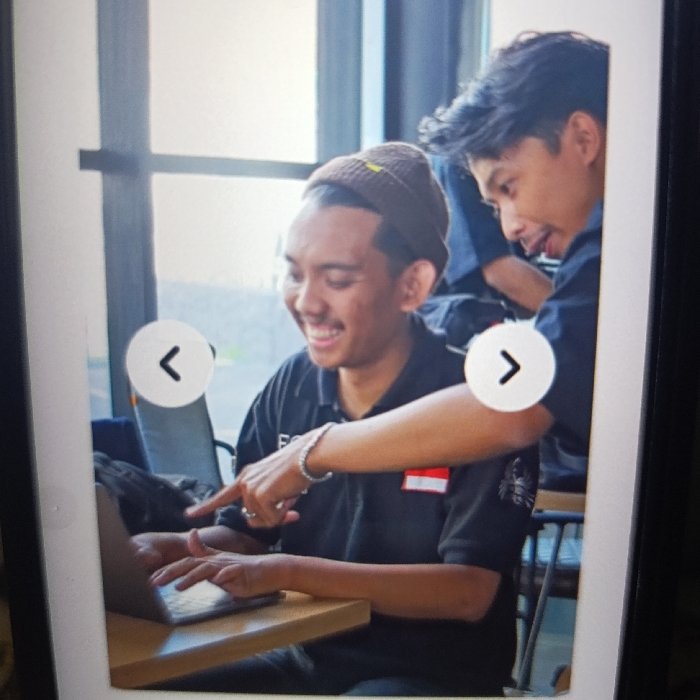The image shows a man wearing a black polo shirt and a brown beanie hat. He is sitting at a wooden table, using a laptop computer. Another man, wearing a dark blue shirt, is sitting behind him, reaching forward with his right hand to point at the laptop screen. The younger man smiles as he looks at the screen. The scene takes place in a cafe setting, with a window visible in the background. The coordinates provided, -7.779 latitude and 114.135 longitude, are within Kecamatan Arjasa, in the East Java region of Indonesia.