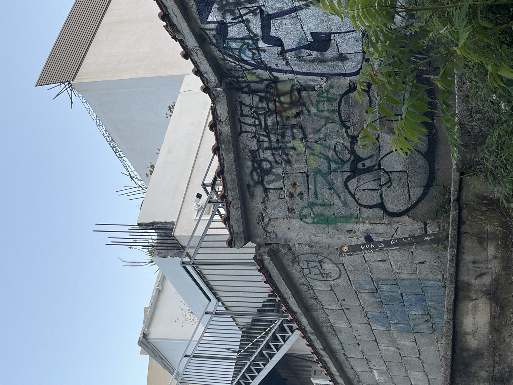 The image depicts an urban scene in Budva, Montenegro, characterized by a mix of architectural elements and graffiti. In the foreground, a weathered concrete wall is covered with various graffiti tags in different colors, including black, green, and blue. A tall, thin, dark-colored post with what appears to be a small skull-like icon at the top is leaning against the wall. Some green foliage is growing near the base of the wall.
Behind the wall, a modern white building with a sloped roofline is visible, topped with exposed rebar. To the left, a metal staircase with railings ascends along the side of a building. Further in the background, another white structure with horizontal siding is seen against a clear blue sky. The overall impression is one of an area with ongoing construction or recent development, juxtaposed with the presence of street art.