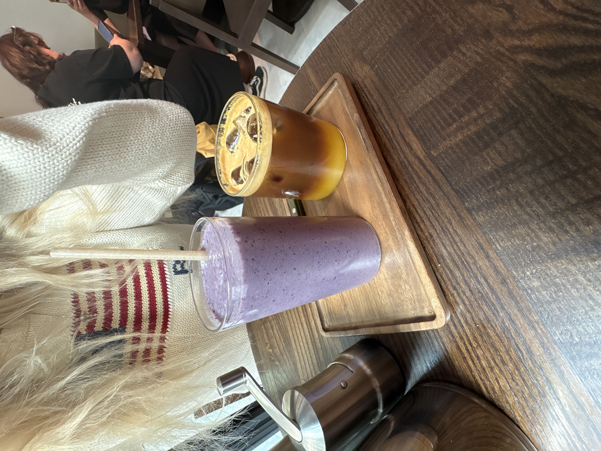 A wooden table holds a wooden tray with two drinks: a purple smoothie with a straw and an iced coffee. A manual coffee grinder is next to the smoothie. An individual wearing a sweater with red and white stripes is seated next to the table. Another person wearing black is seated at the adjacent table, occupied with a cell phone. The photo was likely taken in Milan, Italy, due to the coffee grinder on the table.