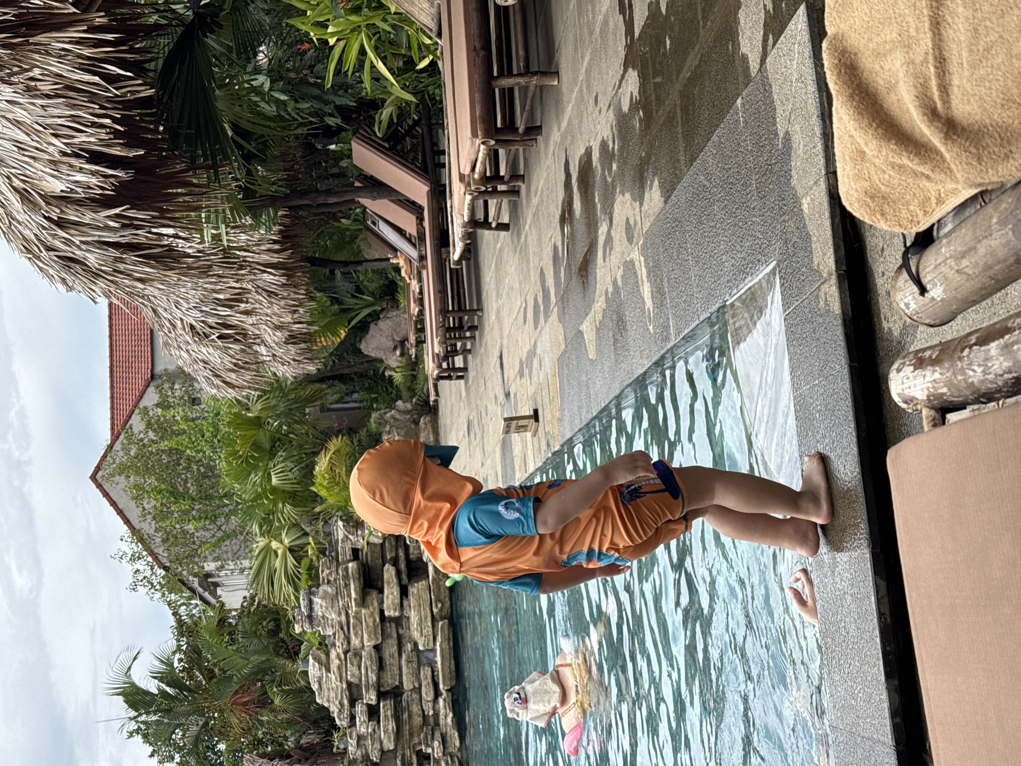 A young child wearing an orange sunsuit and hat stands at the edge of a swimming pool, looking down into the water. In the pool, a person wearing a sun hat and a pink float is visible. The poolside is paved with stone and lined with lounge chairs under a thatched roof structure. In the background, there is a stone waterfall feature and lush tropical vegetation, as well as a building with a red tile roof. The scene appears to be a resort or hotel in Nam Hoa Lư Ward, Vietnam.