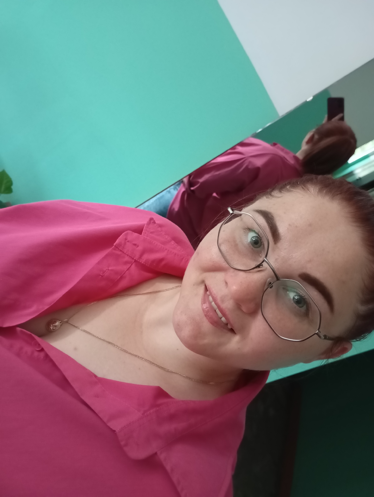 A young woman with light skin, brown hair pulled back in a bun, and green eyes is taking a selfie in a room with teal walls and a reflective surface. She is wearing a bright pink collared shirt and a delicate gold necklace with a circular pendant. She is also wearing hexagonal-shaped eyeglasses. In the reflective surface, a partial reflection of the woman shows her holding a dark-colored smartphone to take the picture. The environment appears to be indoors, with no clear indication of the city of Chaikovsky, Russia, or any other specific location.