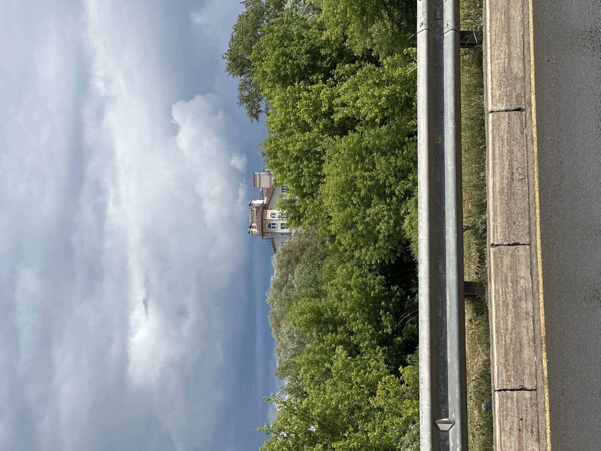 A cloudy, overcast sky looms over a dense line of lush green trees. Partially visible above the trees is a multi-story building with a light-colored facade, adorned with architectural details and several chimneys on its roof. In the foreground, a weathered metal guardrail separates the viewer from a cracked concrete barrier and an asphalt road marked with a yellow line. The scene appears to be a view from a roadside, possibly along a highway or main road on the outskirts of the city of Vladimir, Russia, given the presence of urban architecture peeking through the natural landscape. There are no people or animals visible in the image.
