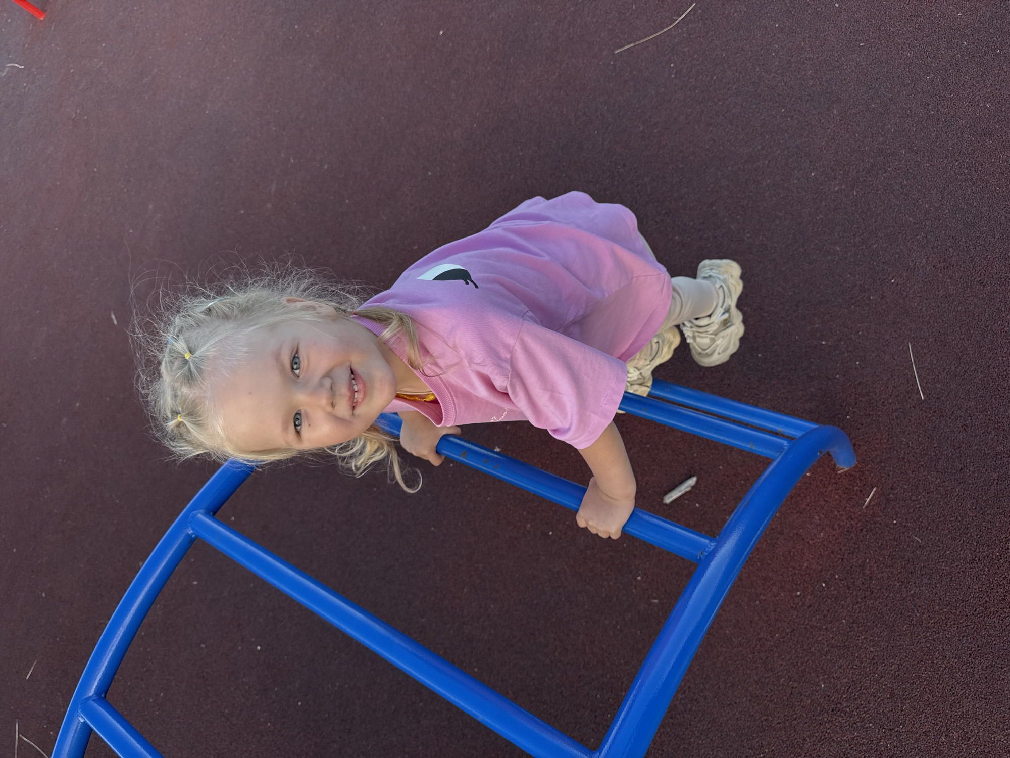A young girl with blonde hair tied in pigtails is on a playground. She is wearing a pink t-shirt with a black graphic and grey pants, along with white sneakers. The girl is lying on a blue monkey bar structure, with her hands gripping the bars. She is smiling at the camera, appearing happy and engaged. The playground surface is a dark, textured material, possibly rubberized, common in modern playgrounds. The background is a blur of the same dark surface, suggesting an outdoor setting. The image appears to be taken during the daytime, with natural light illuminating the scene. The girl's pose and expression convey a sense of playfulness and enjoyment of the playground equipment. There is no visible text in the image.