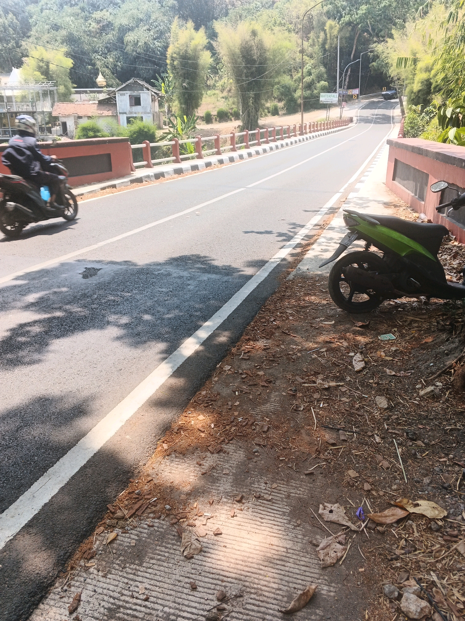 The image depicts a street scene in Semarang, Indonesia, characterized by a paved road winding through a hilly, vegetated landscape. A dark-colored motorcycle with a rider wearing a helmet is traveling on the left side of the road. To the right of the road, a green scooter is parked on a dirt and leaf-strewn shoulder. The road is lined with a red and white barrier and a sidewalk. In the background, there are buildings, bamboo trees, and a dense forest. A sign for "WATERFALL" is visible on the left side of the road. The lighting suggests it is daytime, with shadows cast across the road.