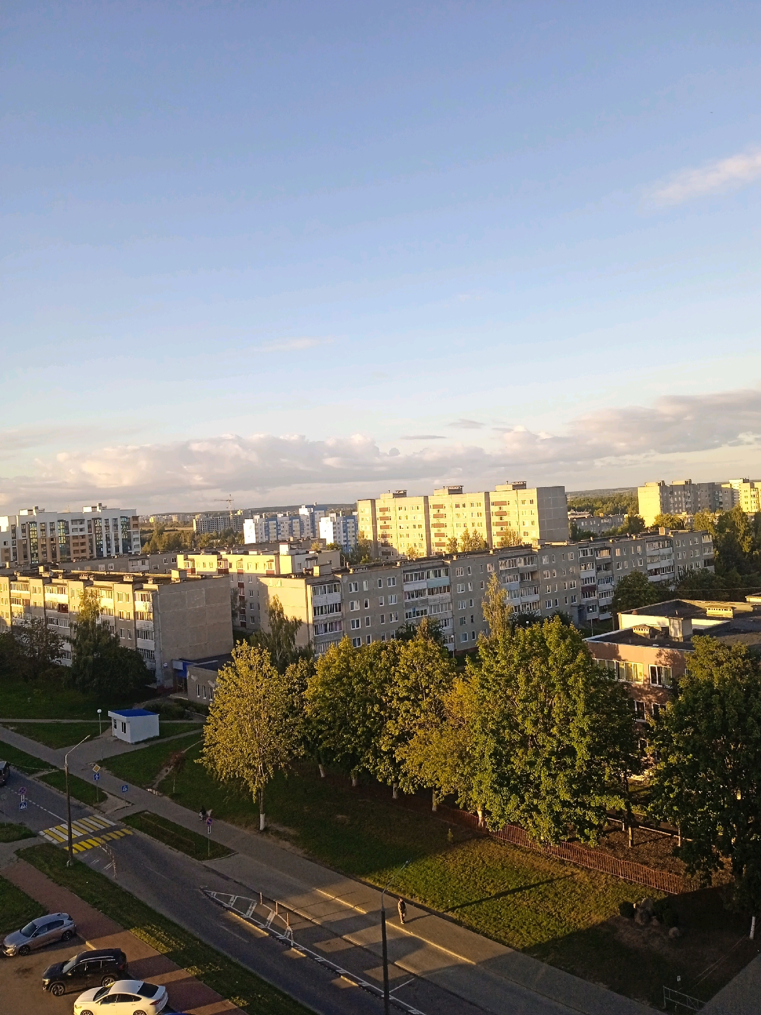 The image captures an aerial view of a residential area in Lida, Belarus, under a clear, pale blue sky with scattered clouds. The scene is dominated by multi-story apartment buildings, characteristic of Soviet-era architecture, with their grey and light-colored facades. Some buildings are illuminated by the warm, late afternoon sun, casting long shadows across the landscape. Trees, predominantly deciduous and green, are interspersed throughout the urban environment, some towering over the lower buildings.
In the foreground, a paved road winds through the area, marked with yellow pedestrian crossings and traffic signs. Several cars are parked along the road or driving on it: a silver sedan, a black SUV, and a white sedan. A lone individual is walking on the sidewalk adjacent to a grassy area. A small, white utility building with a blue roof is situated near the road. The overall impression is that of a typical, lived-in urban neighborhood during a calm, sunny day.