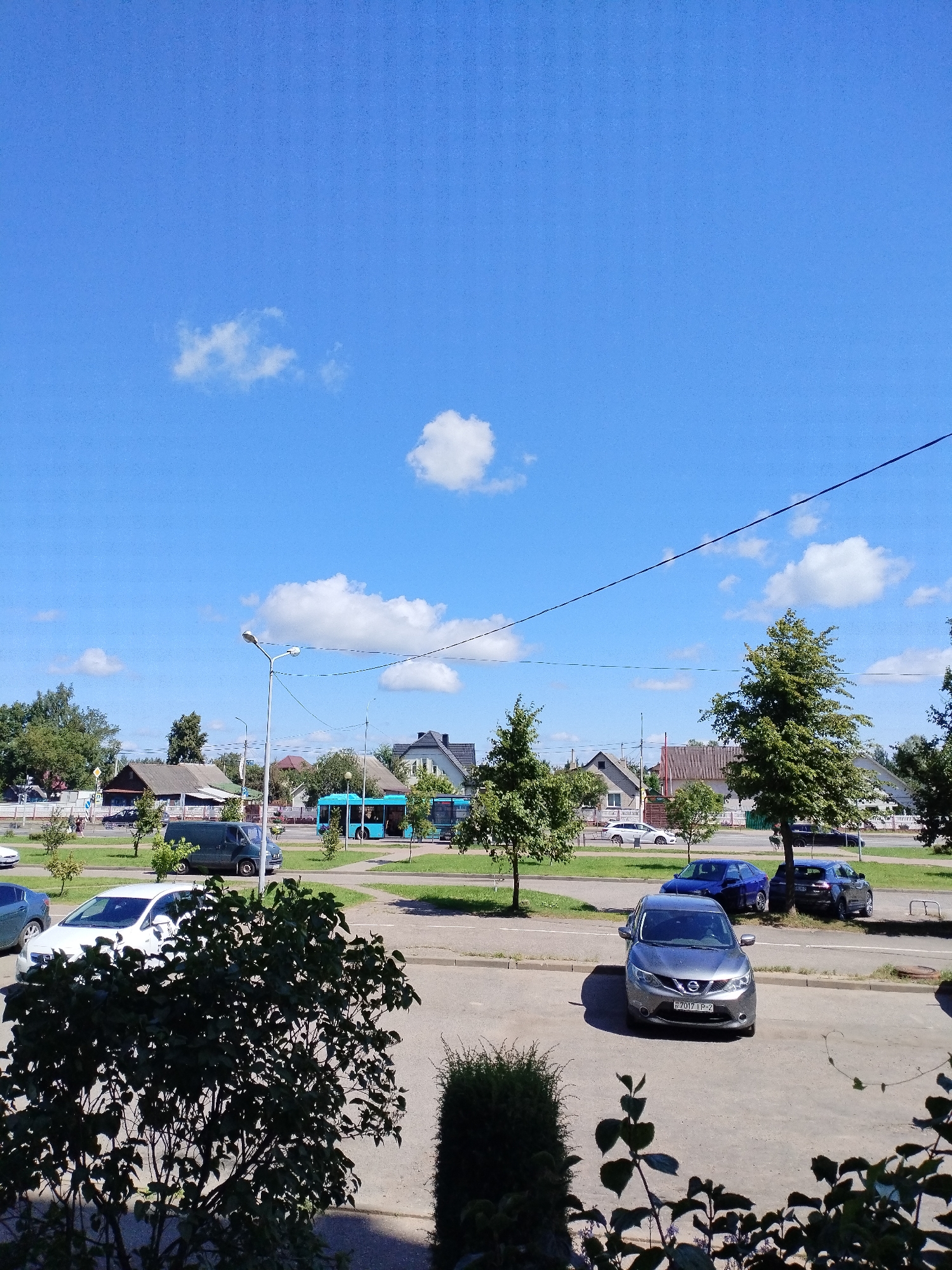 This image captures a street scene in Polatsk, Belarus on a clear day with a bright blue sky dotted with scattered white clouds. In the foreground, a dark-leaved bush partially obscures a concrete surface. Moving further back, several parked cars are visible, including a grey Nissan sedan with a license plate reading "H701TP-2", a dark blue hatchback, and a dark-colored SUV parked behind it. A white sedan is parked on the other side of the street.
The middle ground features a road with a blue bus in motion. Alongside the road are trees and more houses, characteristic of a residential area. A street lamp stands prominently near the center of the image. The background displays more houses with visible roofs and windows, as well as a line of trees. The overall impression is of a typical suburban or town streetscape.