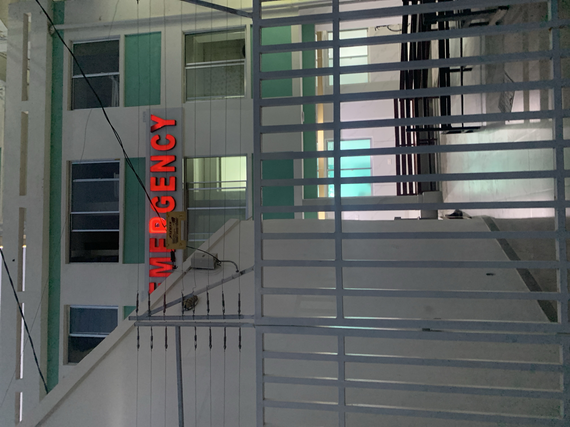 A sideways shot shows the outside of an emergency room. The emergency room sign has red lettering. It is an older white building with green columns. A wire fence hides some of the building. The background is dark. Based on the style of architecture, it is likely located in Dar es-Salaam, Tanzania.