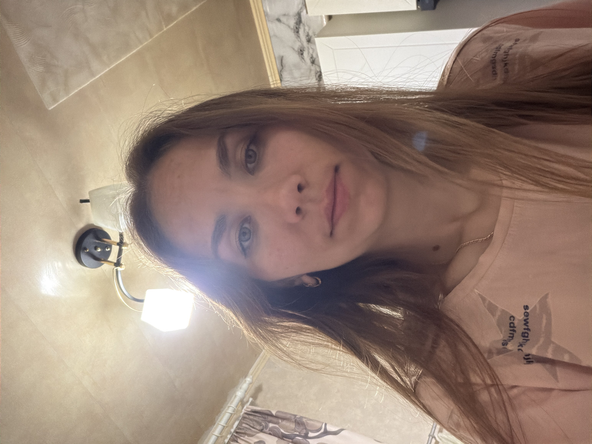 The media file contains a close-up, self-taken portrait of a young woman with long, light brown hair. She is looking directly at the camera. Her eyes are blue-gray, and she has a light complexion with some freckles visible. She is wearing a light pink t-shirt with a star graphic and some text on it. The text is partially visible and appears to read "sewfgthjk calm." She is wearing small hoop earrings.
The setting appears to be an indoor location, possibly a bathroom or a hallway, with beige tiled walls and a wall-mounted light fixture. The light fixture has a black base and a white cube-shaped shade that is illuminated. The lighting in the scene is bright, casting shadows and creating a glare on the woman's hair and face. The overall impression is informal and candid. There are no clear indicators of the time of day or weather conditions. The location context provided is Tambov, Russia, but this cannot be visually confirmed from the image itself.