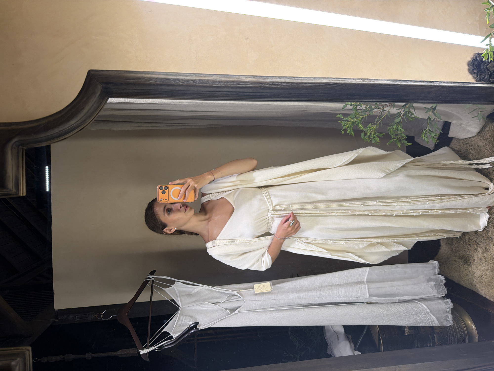 A woman is captured in a mirror selfie, standing in what appears to be a boutique or fitting room. She is wearing an elegant, cream-colored, flowing dress with intricate rope or bead detailing around the bodice and waist, and possibly extending to the shoulders. The dress has a relaxed, artisanal aesthetic. She holds an orange smartphone with a textured case to take the photo, her gaze directed towards the screen. A delicate bracelet is visible on her left wrist, and a ring adorns her right ring finger.
The mirror she is using has a substantial, dark, possibly carved wooden frame, reflecting the room around her. Reflected in the upper portion of the mirror is a light beige wall and a long, horizontal light fixture. To the upper right, some green foliage, likely decorative, is visible. Below the woman's reflection, another light-colored garment, possibly a linen or sheer fabric dress, hangs on a wooden hanger, with a small, unreadable tag attached, further indicating a retail environment. The background reflected behind this hanging garment is dark and indistinct.
The overall aesthetic, featuring natural textures, flowing fabrics, and a rustic mirror, is consistent with a craft-focused boutique or studio, fitting the provided location context of the Ubud District, Indonesia. The scene is illuminated by artificial light, suggesting an indoor setting.