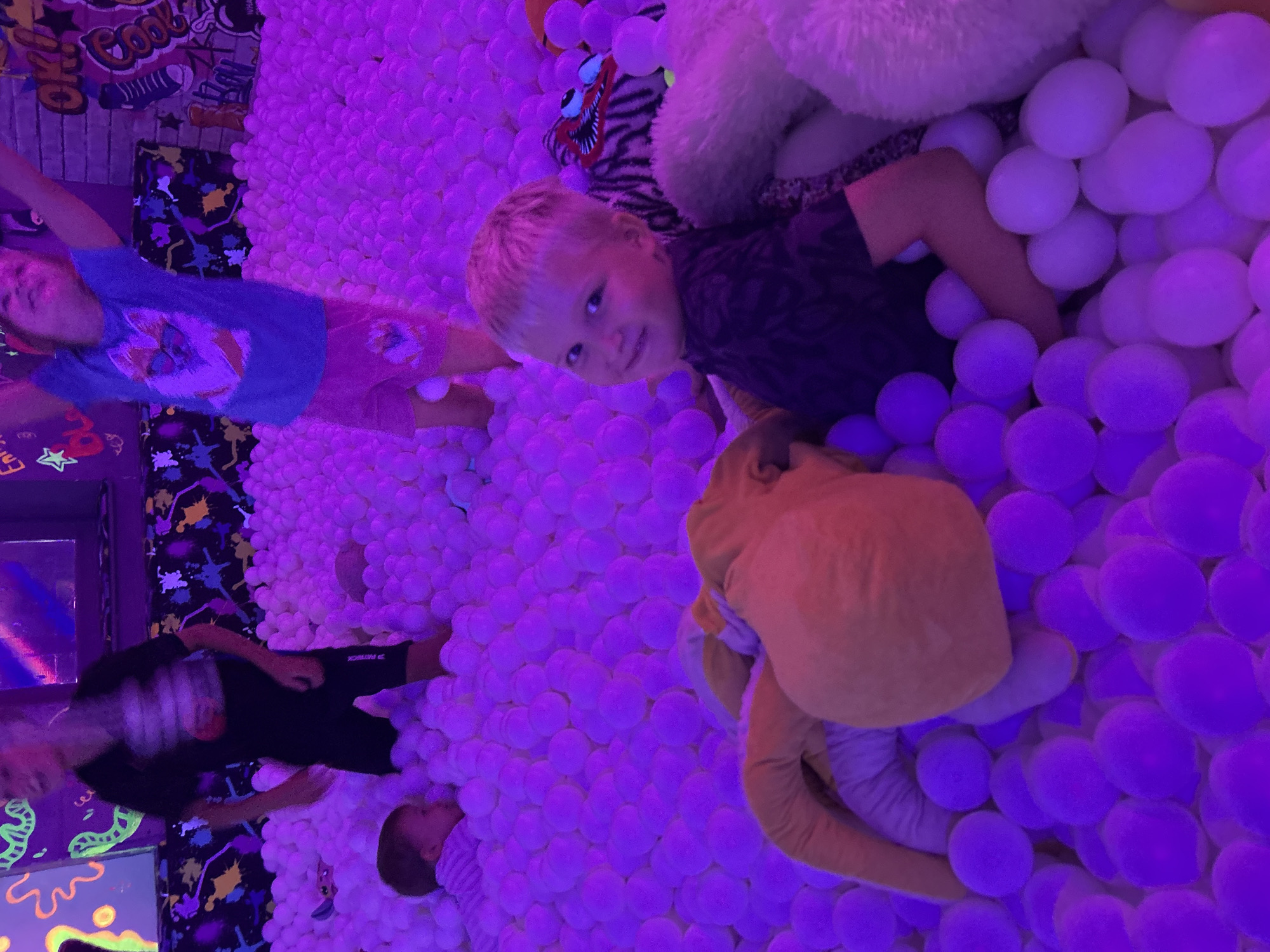The image captures a vibrant indoor scene, likely a play area with a ball pit, bathed in purple neon lighting. In the foreground, a young Caucasian boy with blond hair and blue eyes smiles at the camera, nestled among white plastic balls and plush toys. Behind him, a stuffed animal with tiger-like patterns and a large, fluffy white stuffed bear are visible. The background features two other children, one a toddler lying down in the ball pit and another, an older child, standing and actively playing. The walls are adorned with colorful, abstract graffiti art and neon graphics, suggesting a lively entertainment venue. The overall atmosphere is one of playful energy and childhood fun. This scene is not located in the city of Chaikovsky, Russia.