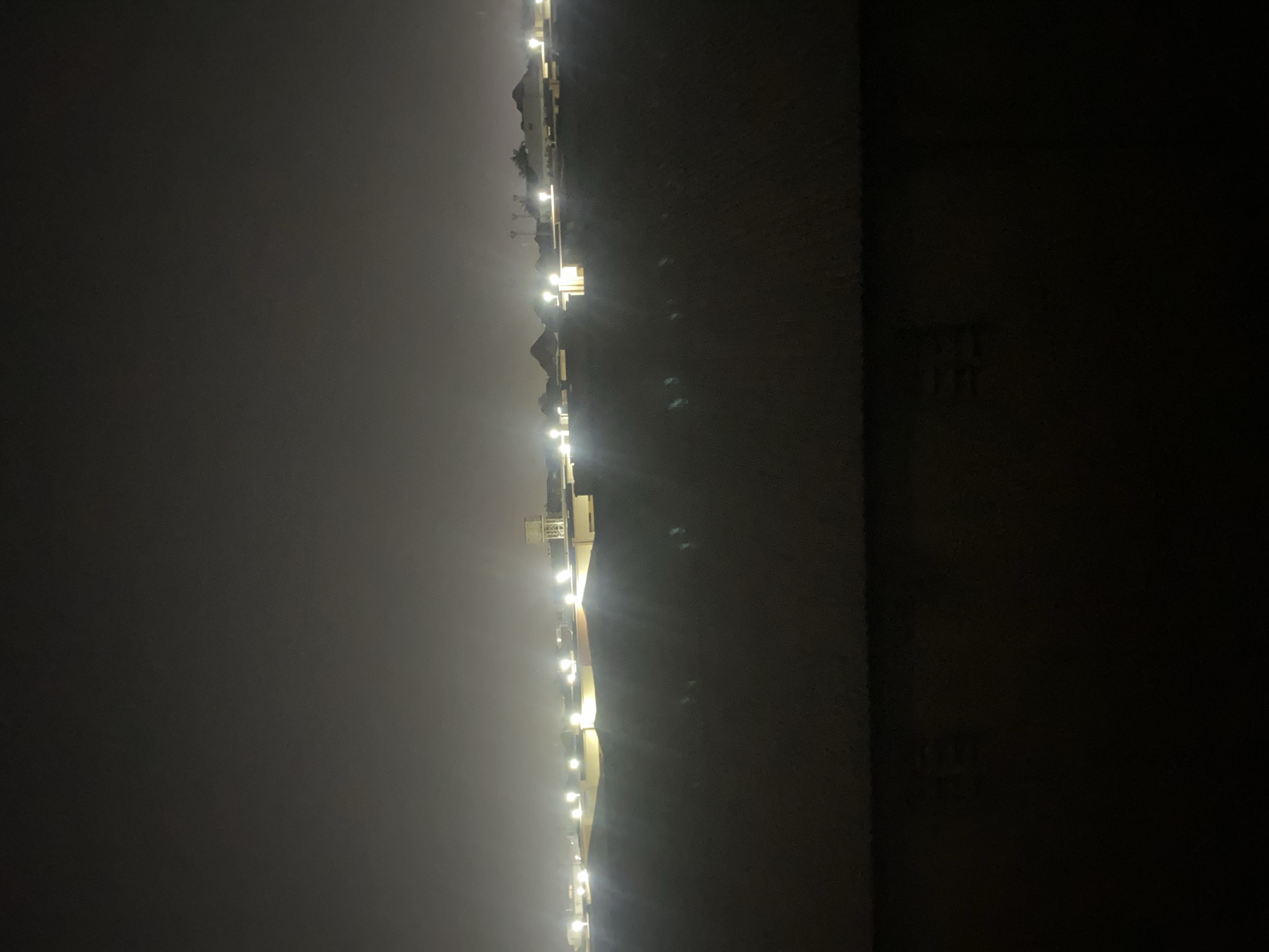 The image depicts a nighttime view of an urban landscape in Fagge A, Nigeria. The foreground is dominated by a dark, textured surface, possibly a wall or ground, with subtle hints of reflected light. Beyond this, a row of buildings stretches across the mid-ground, illuminated by numerous bright, scattered lights. These lights appear to be streetlights or lights from within the structures, creating a hazy glow in the dimly lit environment. In the far distance, the silhouette of a water tower is visible, along with the faint outlines of more buildings and possibly some vegetation against the dark, overcast sky. The overall impression is one of a city at night, with artificial lighting cutting through the darkness and illuminating the built environment.