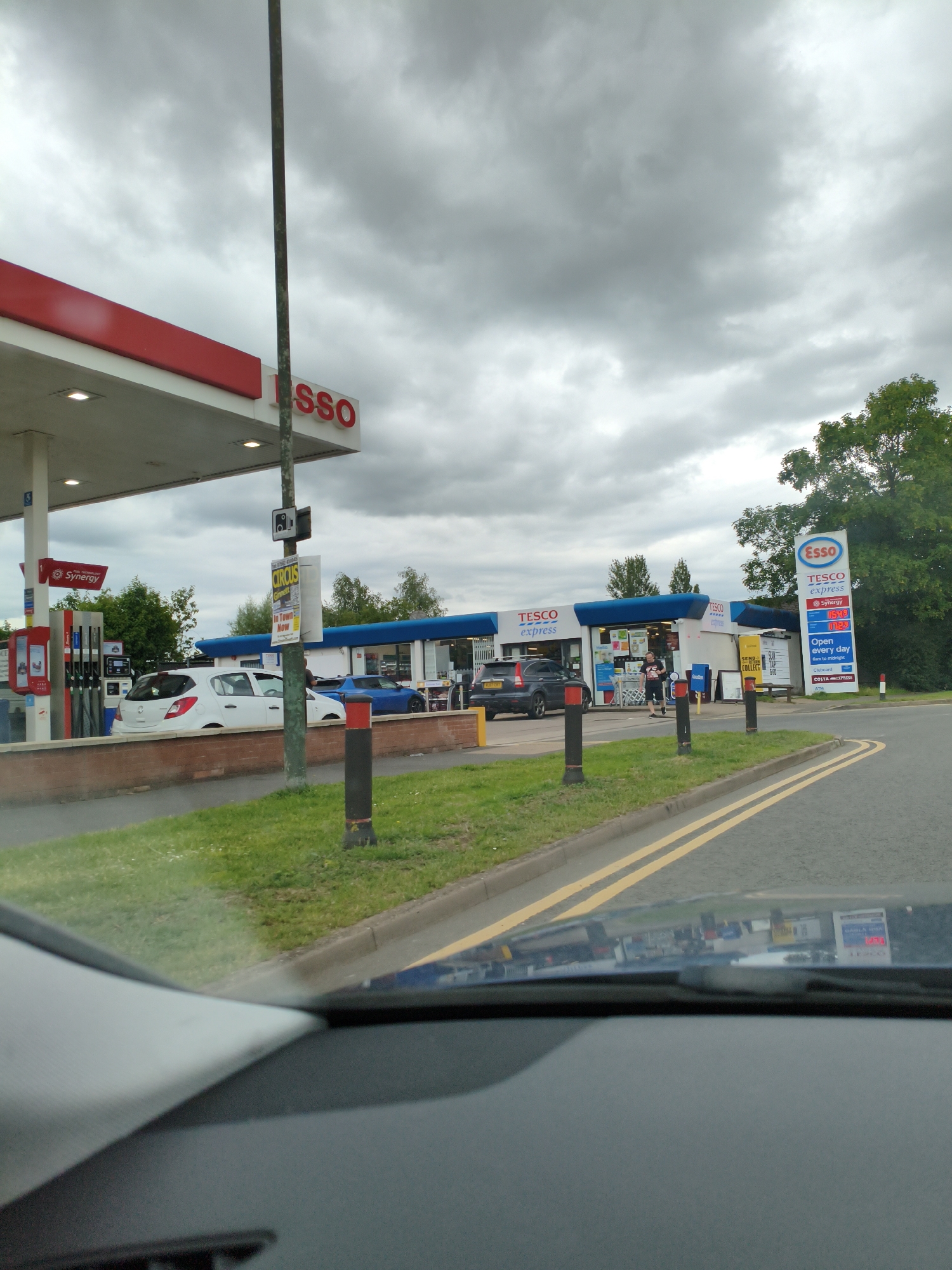 An eye-level, medium shot captures an exterior scene at a Tesco Express petrol station on a cloudy day. The Esso branding is prominent, with a large red and white sign featuring the Esso logo and "Tesco express" text, alongside fuel prices. Several cars are visible, including a white hatchback at a pump, a dark SUV, and a blue sports car. Two individuals, both appearing to be male, are near the entrance of the Tesco Express store. The foreground shows the interior of a vehicle, with the dashboard and windscreen visible, reflecting some of the scene. The environment includes a paved road with yellow lines, a grassy area, and trees in the background. The location is identified as Wychavon, United Kingdom.