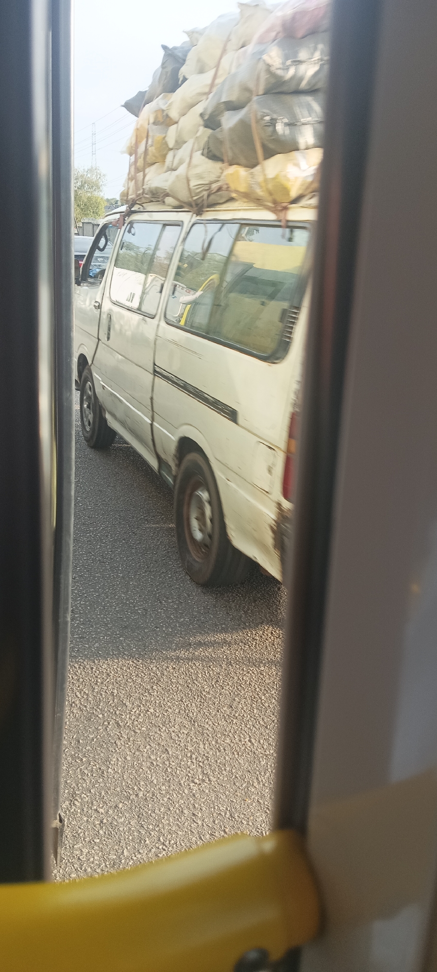 The image displays a white, well-worn van, possibly a minivan or small bus, captured from the perspective of an adjacent vehicle. The van is heavily loaded with numerous large, light-colored and grey sacks piled high on its roof, secured with ropes. This cargo appears to extend significantly above the vehicle's normal height.
The van is situated on an asphalt road, suggesting it is either stationary in traffic or moving slowly alongside the photographer's vehicle. Signs of wear and tear, including dirt and some rust, are visible on the van's body, particularly near the rear wheel and taillight. Horizontal dark stripes are present on its side panels. Through the van's windows, reflections show parts of the surrounding environment, including what might be another vehicle.
In the background, under a bright, clear sky, faint outlines of power lines or utility poles and some sparse foliage are visible. The scene takes place during daylight, likely on a sunny day. The photograph is taken from inside another vehicle, with parts of its interior, such as a yellow handle and vertical frame elements, partially framing the view. This type of heavily laden transport is a characteristic sight in many regions, including Gura Top, Nigeria, where the image was reportedly taken. No visible text is discernible on the van or its cargo.