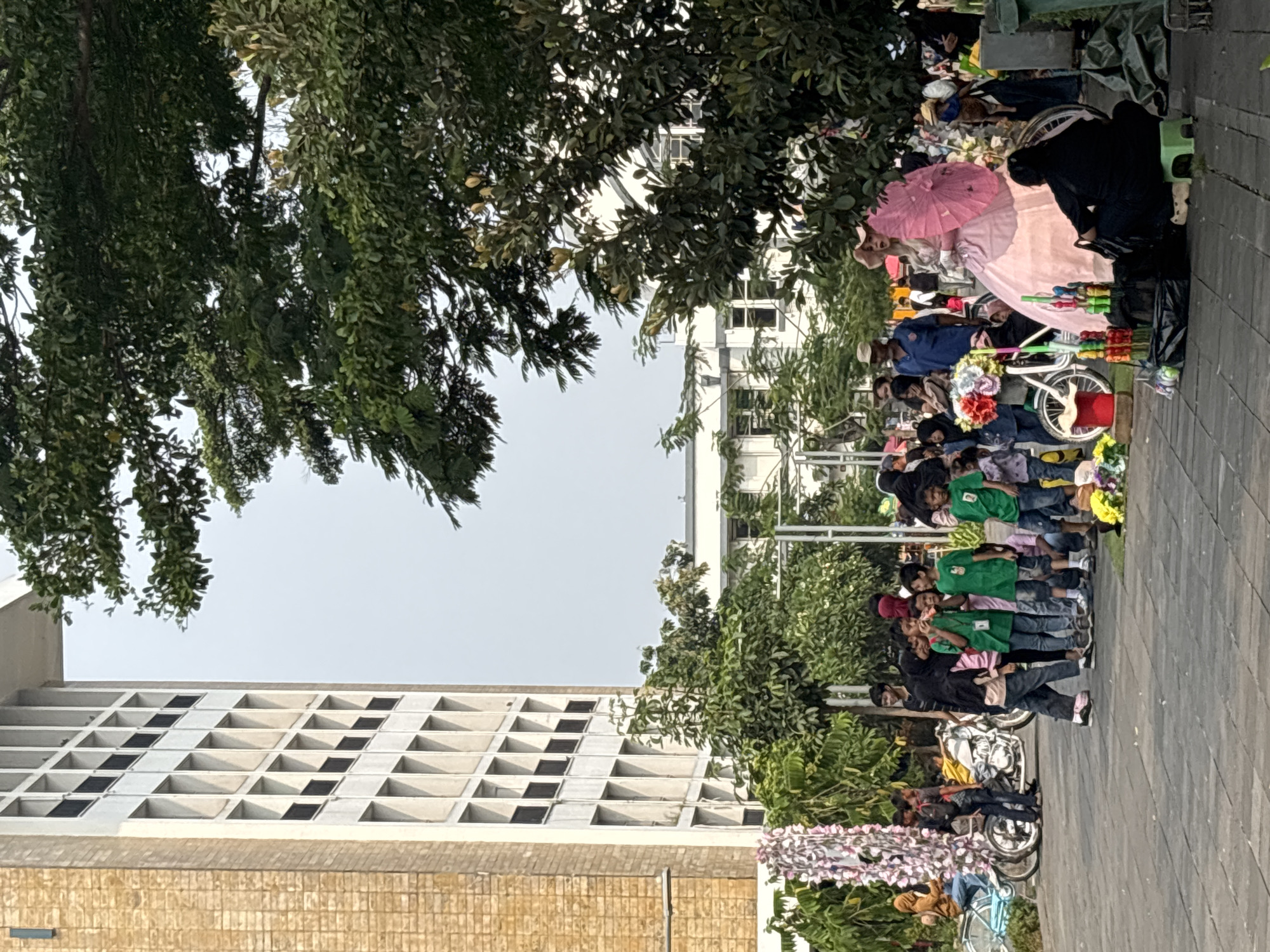 The image depicts a scene in a public outdoor area with a tall, beige building with many windows on the left. In the foreground and midground, there is a gathering of people, many of whom appear to be children, some wearing green t-shirts. There are also adults present. Several objects are visible, including bicycles, motorcycles, colorful flowers arranged as decorations, and umbrellas. A person dressed in a large pink gown and hat, holding a pink umbrella, is a prominent figure in the center. In the background, more trees and buildings can be seen. The overall atmosphere suggests a public event or gathering, possibly a market or festival. While the specific location within West Jakarta, Indonesia, is not explicitly stated in the image, the presence of a large building and an outdoor gathering area is consistent with urban environments in that region.