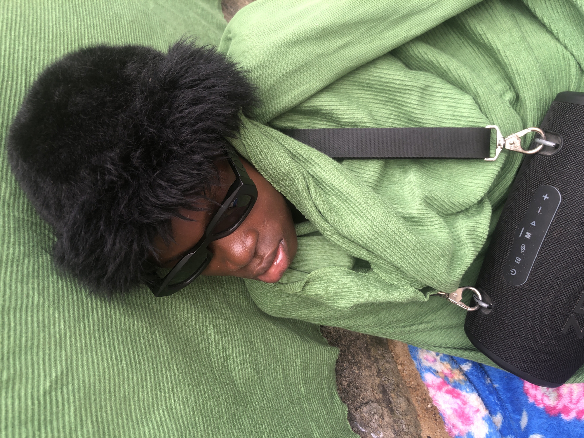 A person wearing a black fur hat and black sunglasses is lying down on a green corduroy fabric. The person is draped in more green corduroy fabric, and a black portable speaker with a strap is hanging from the fabric. A colorful blanket is visible underneath the green fabric on the left side of the image. The scene appears to be outdoors, as suggested by the rough, rocky ground beneath the blanket. The lighting indicates it is daytime. The overall impression is one of relaxation or rest. The image is a close-up shot, focusing on the person and their immediate surroundings. The specific location in Ado, Nigeria, cannot be determined from the visual information alone.