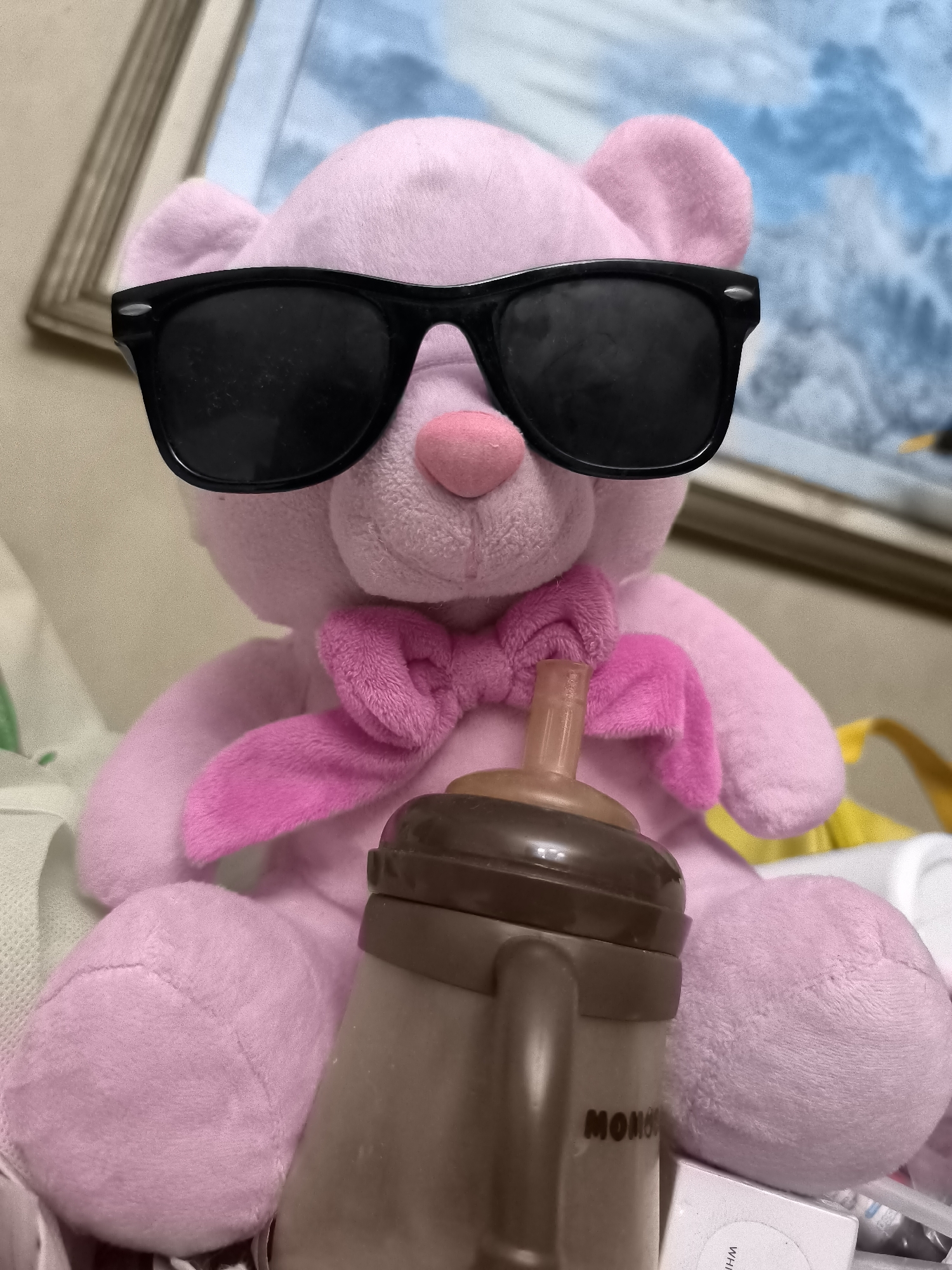 A pink teddy bear wearing black sunglasses and a pink bow tie is sitting in an upright position. It is holding a brown baby bottle with a light brown nipple. The teddy bear is positioned in front of a framed painting. The environment appears to be indoors, possibly a room. The image does not provide any information about the location in North Jakarta, Indonesia.