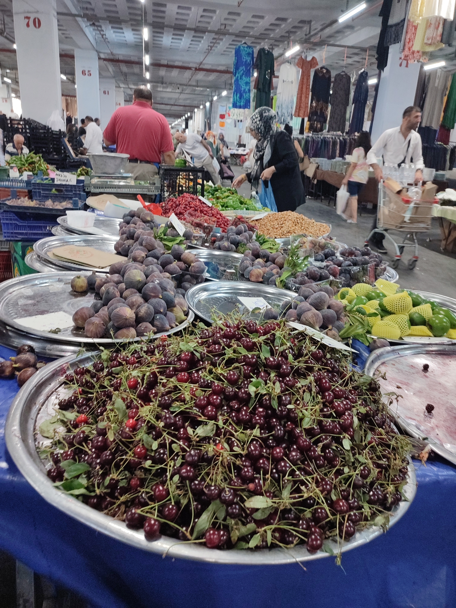 The image depicts a bustling indoor market, likely in Kadıköy, Turkey, with a focus on a fruit stall. In the foreground, a large silver platter is overflowing with dark red cherries, their stems and green leaves still attached, creating a vibrant display. To the left and behind the cherries, another silver platter holds a mound of ripe figs, their purple skins glistening. Further back, more fruit is visible, including what appear to be green apples or pears, some nestled in yellow protective netting, and piles of what looks like dried nuts or seeds.
In the mid-ground, a man in a red shirt is visible behind a scale, suggesting he is a vendor. Shoppers are moving through the market. A woman wearing a headscarf browses produce, and a man with a shopping cart is seen further down the aisle. The market environment is suggested by overhead lighting, clothing racks displaying various garments in the background, and structural elements like pillars marked with numbers (70 and 65 are visible). The scene is well-lit, capturing the lively atmosphere of a local marketplace.