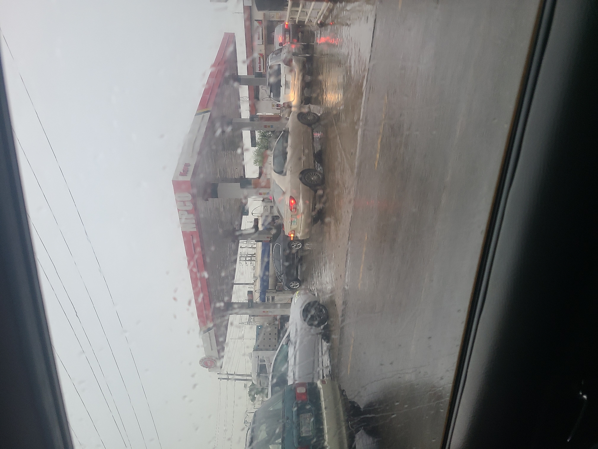 This image depicts a gas station scene in Karu, Nigeria, during what appears to be a rainy day. The perspective is from inside a vehicle, looking out through a wet window, with raindrops visible on the glass. Several cars are lined up at the gas pumps under a large, red and white canopy. The ground is wet and reflective, indicating recent or ongoing rain.
The most prominent object is the gas station structure with its canopy and pumps. Cars are the primary subjects, with at least three visible in the queue. The cars are of various colors and models, but none are actively fueling up at this moment. The overall atmosphere is somewhat dreary due to the weather.
The sky appears overcast, suggesting it's daytime, likely late morning or afternoon, though the exact time is difficult to ascertain due to the weather conditions. The presence of cars waiting at a gas station suggests an activity of refueling or perhaps a queue due to limited services or high demand.
Visible text includes "NIPCO" on a sign and what appears to be "Kano" on another sign. The context of Karu, Nigeria, is provided. The image captures a mundane, everyday scene of a gas station under adverse weather conditions, with the rain adding a layer of atmosphere and altering the visual quality of the scene. The focus is on the vehicles and the infrastructure of the gas station, viewed through the slightly obscured lens of a wet car window.