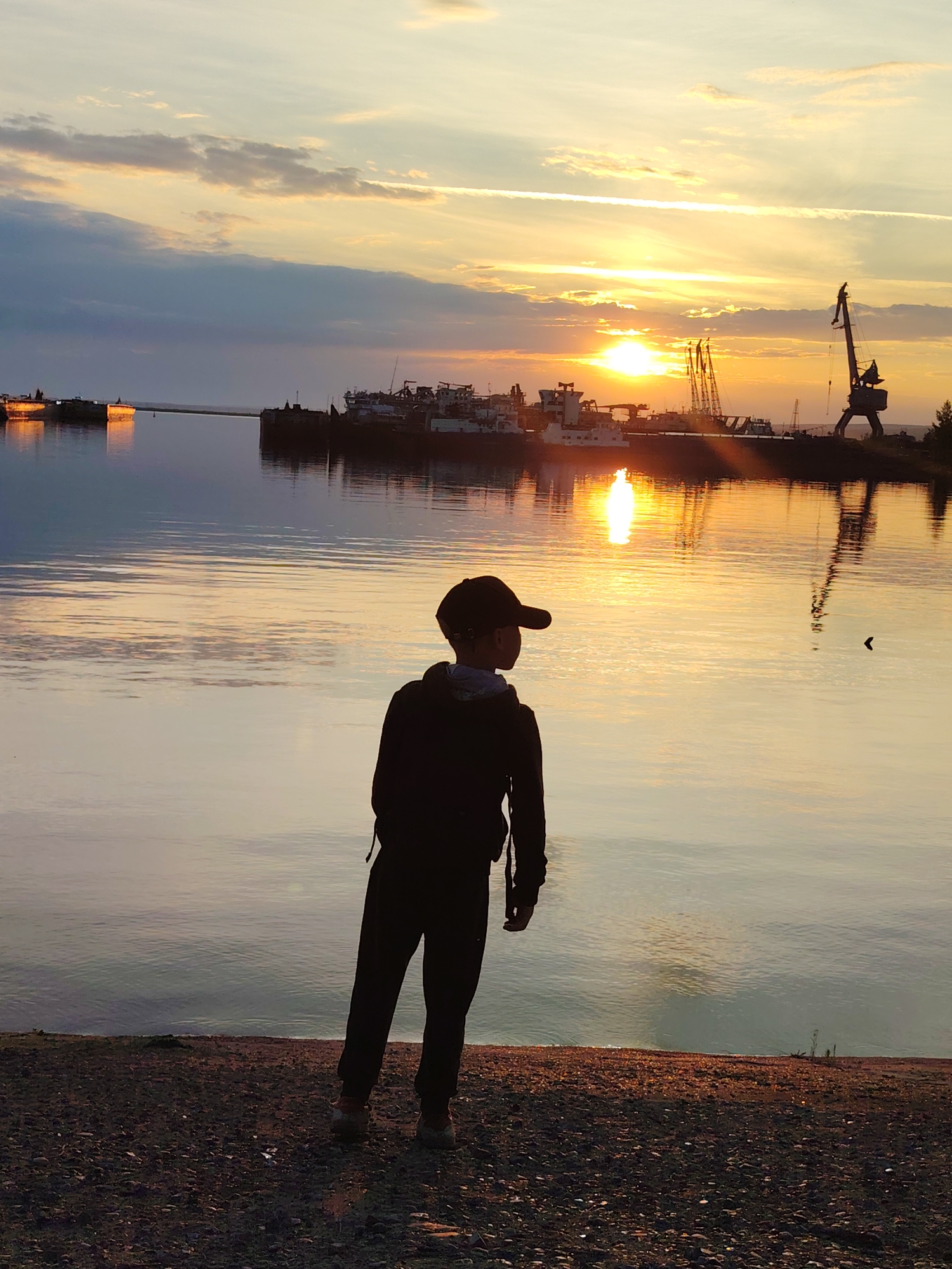 An outdoor, eye-level, vertical, wide shot captures a silhouetted young person facing away from the camera, looking out at a body of water during sunset. The individual is wearing a dark hooded sweatshirt, dark pants, and a baseball cap. Their reflection is visible in the calm water, which is illuminated by the orange and yellow hues of the setting sun.
In the background, a harbor is visible with several docked ships and large cranes. The sky displays a gradient of colors from pale yellow near the horizon to blues and grays higher up, with thin, wispy clouds streaking across. The sun is low on the horizon, casting a bright reflection on the water. The foreground consists of a pebble-strewn shoreline. The scene is likely the Kama River embankment in Chaikovsky, Russia, given the presence of a harbor, ships, and cranes, characteristic of the city's industrial and riverside setting.