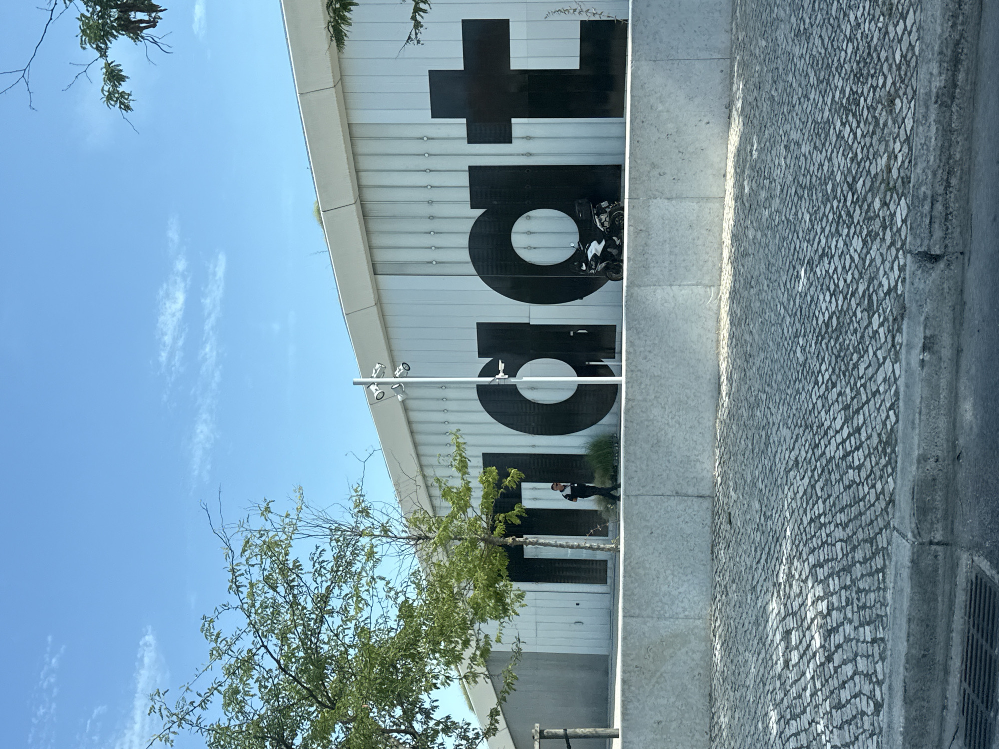 The image captures a frontal view of the MAAT (Museum of Art, Architecture, and Technology) building in Lisbon, Portugal, on a clear day. The museum's facade is characterized by large, black sans-serif letters spelling out "MAAT". The building itself features a white, corrugated metal exterior.
In front of the building, a person in dark clothing is walking towards the right side of the frame. To the right of this person and in front of the "a" and "t" of "MAAT", a white and black motorcycle is parked.
The foreground of the image is dominated by a cobblestone street, with a concrete curb and a metal drain grate visible at the bottom. A low, grey concrete wall runs horizontally across the middle of the frame, separating the street from the museum's entrance area. Trees with green foliage are present on the left and right sides of the image, framing the view of the building. The sky is bright blue with a few wispy clouds.