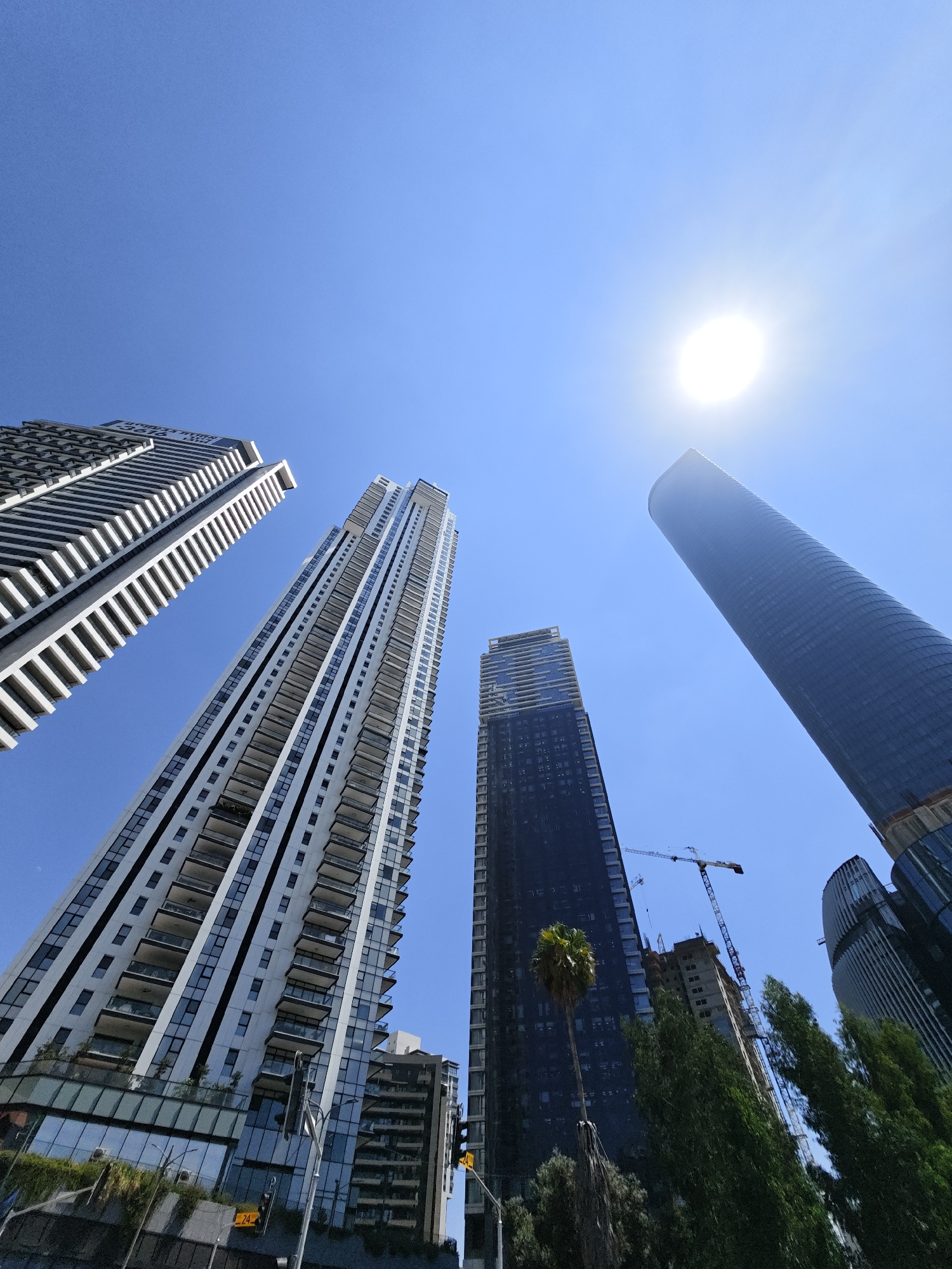 The image shows a low-angle view of several tall modern buildings against a clear blue sky, with the sun prominently visible and partially obscured by a haze. The architecture is predominantly glass and concrete, with visible balconies on some of the residential towers. Construction cranes are present, indicating ongoing development. Trees and foliage are visible at the base of some of the buildings. The scene suggests an urban environment in a sunny climate. Based on the context provided, this location is Givatayim, Israel.