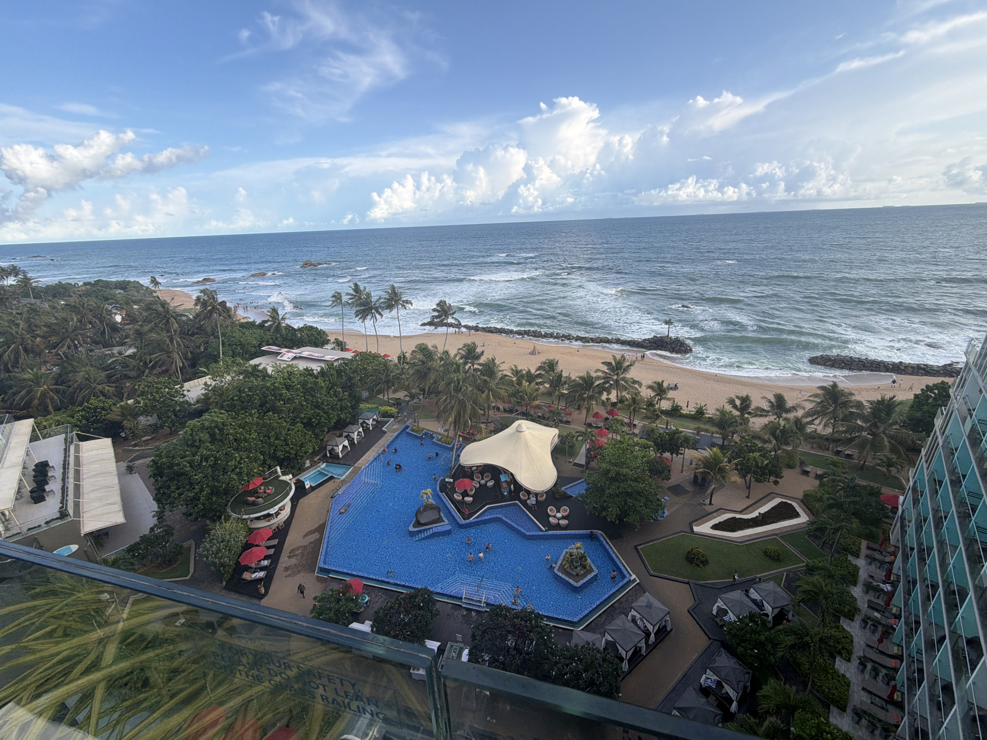 The image captures a high-angle view of a resort area adjacent to the ocean, likely taken from a tall building or hotel room in Galle, Sri Lanka. The scene features a large, irregularly-shaped swimming pool with blue tiles, surrounded by sun loungers, palm trees, and various structures, including cabanas and dining areas with red umbrellas. People are seen swimming in the pool and walking around the resort grounds. Beyond the resort, there is a sandy beach with palm trees and the ocean, with visible waves and a cloudy sky above. The ocean appears calm, and the sky is partly cloudy. Some buildings and vegetation are present in the background, and a portion of the building from which the photo was taken is visible in the foreground, showing the glass railing of a balcony.