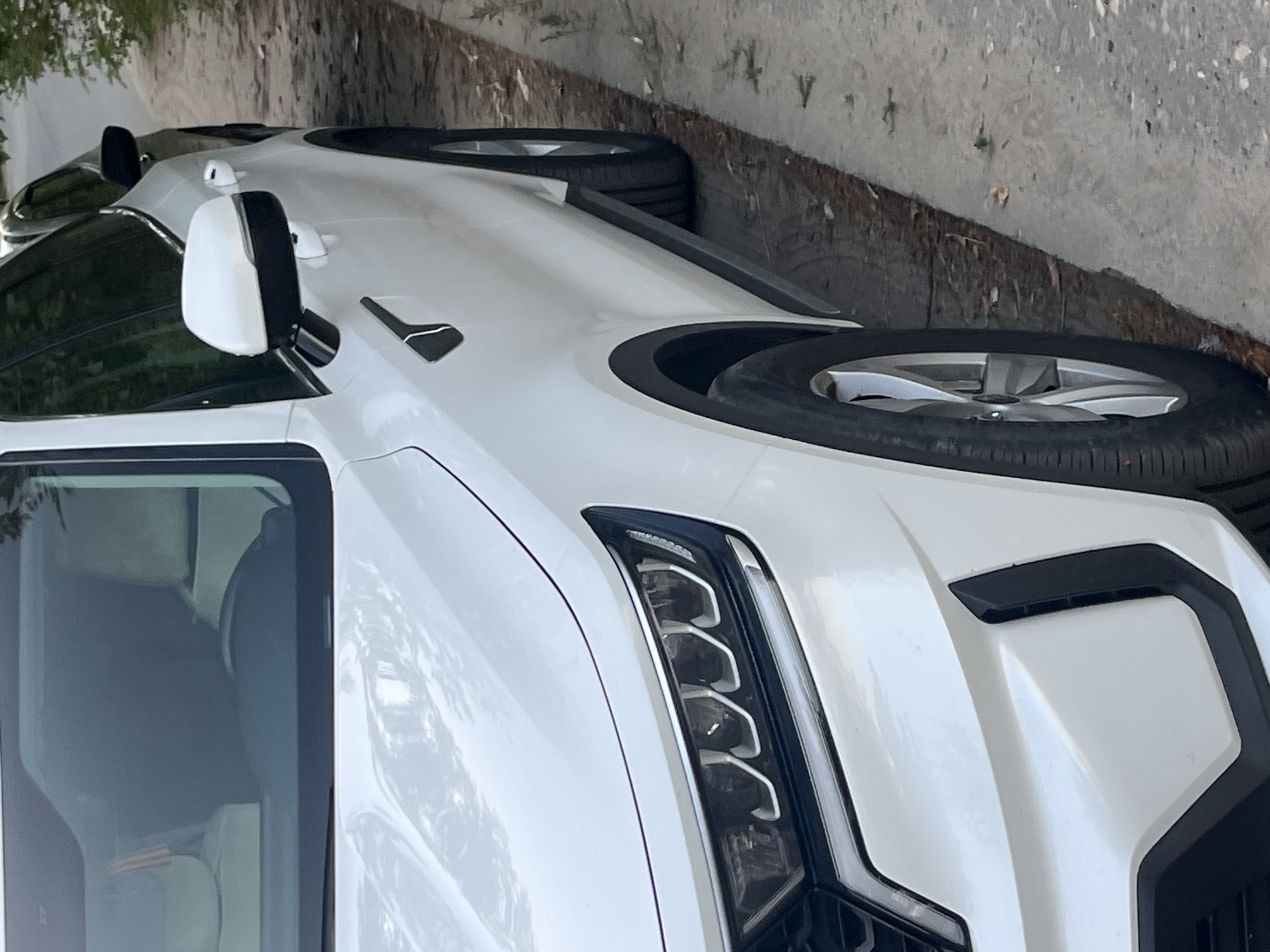 A white car is parked on the side of a road in Volgograd, Russia. The car is a modern SUV with sleek headlights and a black trim around the wheel wells. The car is parked on a concrete curb next to a dirt shoulder with some scattered leaves and small plants. The sun is shining, casting shadows on the ground. In the background, another car is visible, parked behind the white SUV. The environment appears to be a quiet street, possibly in a residential area or on the outskirts of the city.