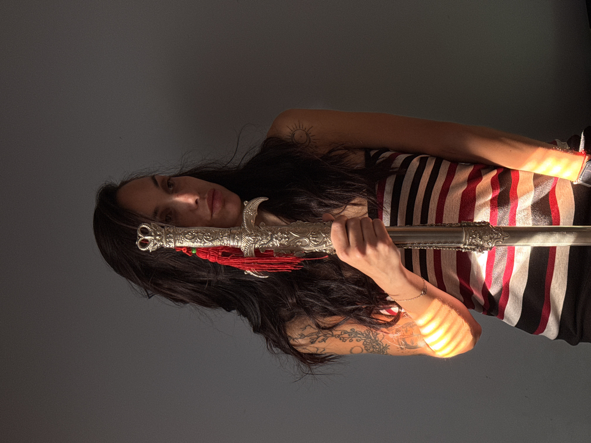 A portrait photograph captures a woman holding an ornate sword horizontally across her body. She has long, dark hair that frames her face and flows over her shoulders. Her expression is neutral, and she gazes directly at the viewer. She is wearing a striped top with red, white, and black vertical stripes, paired with dark-colored bottoms.
Several tattoos are visible on her arms: a circular sun and moon design is inked on her left shoulder, and intricate floral or vine patterns adorn her right arm. The sword she holds features a highly decorative, silver-colored hilt and scabbard, embellished with detailed scrollwork and red tassels near the hilt.
The scene is set indoors against a plain, dark grey wall, suggesting a simple background. Strong natural light, likely sunlight streaming through a window with horizontal blinds, illuminates the right side of her body and the sword. This light creates distinct, parallel shadow lines across her right arm and torso, indicating a sunny day, likely in the late morning or afternoon. The left side of her face and hair are partially shadowed, creating a high-contrast lighting effect. There is no visible text in the image.