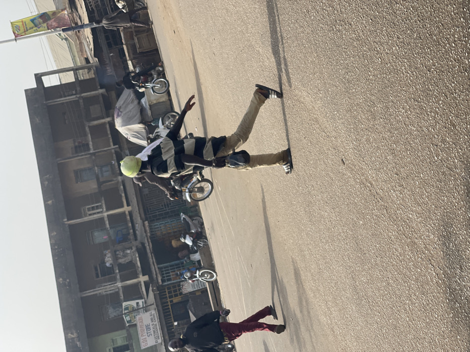 The scene depicts a sunlit street in Chikaji, Nigeria. A male subject is observed in the mid-ground, walking from the right to the left across a paved road. He wears a light-green head covering, a horizontally striped t-shirt (dark and light bands), light-colored trousers, and dark slides with white stripes. He carries a dark, possibly plastic, bag in his left hand, and his right arm is extended laterally. To the left of this individual, another male subject walks from left to right, dressed in a dark long-sleeved shirt and red trousers.
In the background, a two-story building with a weathered facade features multiple windows on its upper floor and several commercial shops at ground level. One visible shop sign reads "GDI PROVISION STORE," with additional text "ELECTRICALS AND ALL RECHARGE CARDS." Several motorcycles are present in the mid-ground and background; some are parked near the shops, and others appear to be in motion or stationary with riders. A light-colored canopy is visible further right in the background, partially obscuring a structure. Another male subject, wearing dark clothing, walks away from the viewer on the far right. A tall, multi-segment pole with a yellow and white banner stands on the right side of the frame. Long, distinct shadows are cast across the asphalt road surface, indicating direct overhead sunlight.