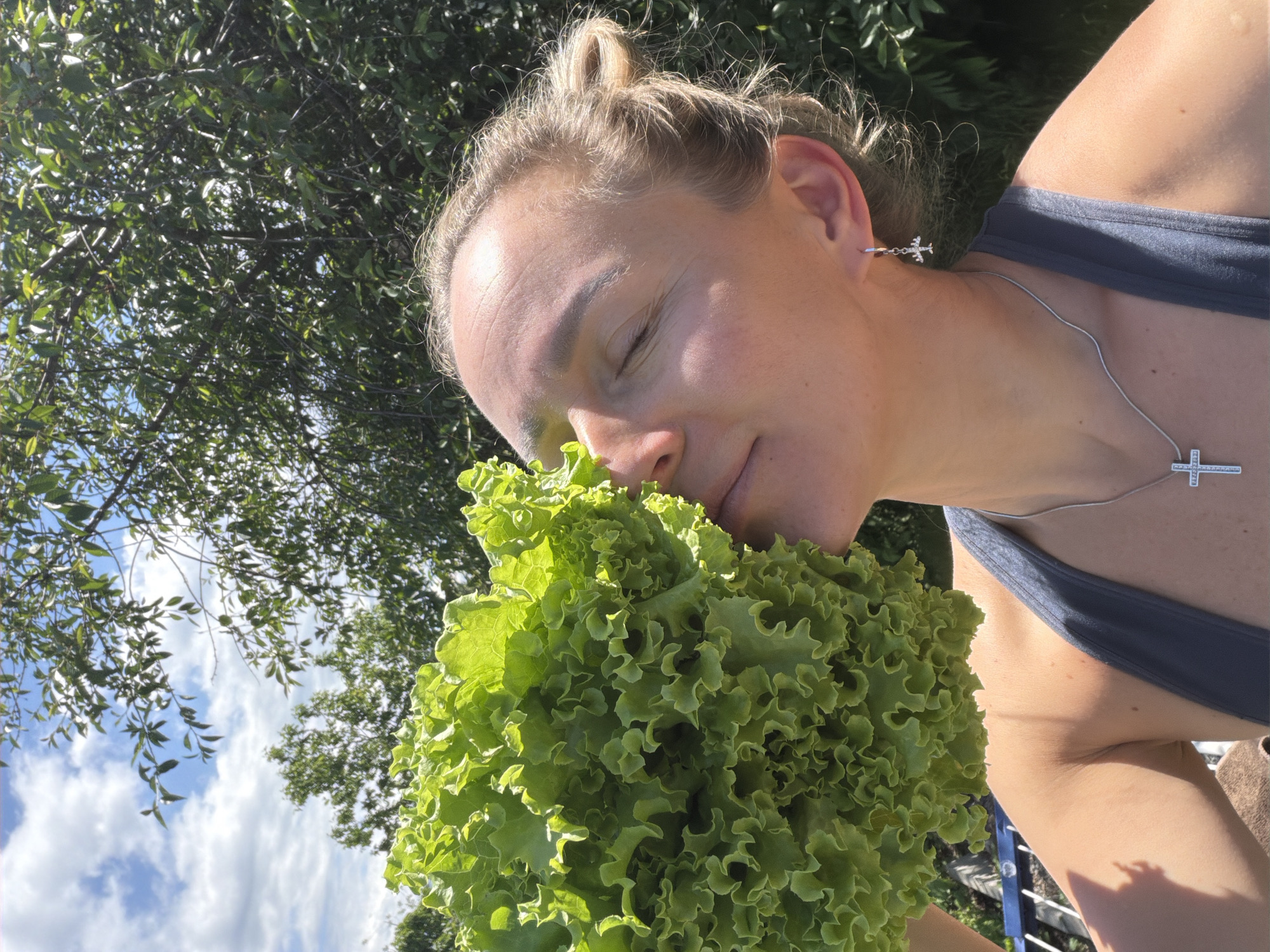 An adult female, with light-colored hair styled in a bun, is depicted outdoors on a sunny day. Her eyes are closed, and she holds a large, vibrant green head of curly leaf lettuce close to her face, with its leaves touching her nose and mouth area. She wears a dark gray tank top, a silver-colored necklace with a studded cross pendant, and a silver-colored hoop earring with an airplane charm in her right ear. Her skin exhibits a tan complexion and some fine lines on her forehead. The background features a bright blue sky with scattered white cumulus clouds and dense green tree foliage, all illuminated by strong sunlight, casting distinct highlights and shadows. A dark railing and a brown object are partially visible in the lower left. The specific location in Chaikovsky, Russia, is not visually identifiable from the image content.