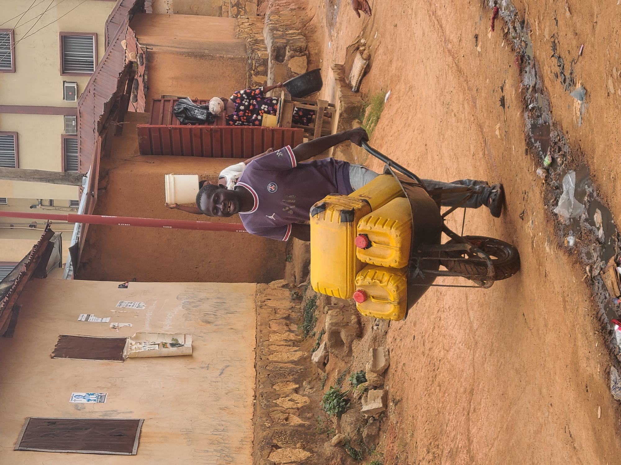 A medium shot captures a man in Jos, Nigeria, pushing a wheelbarrow loaded with three yellow jerrycans. The man is of African descent, wearing a dark purple sports jersey. The wheelbarrow is black and shows signs of wear. The jerrycans appear to be full and have red caps. The surroundings suggest an urban environment with buildings, some dilapidated, lining a dirt road. Debris and signs of a drainage ditch are visible along the side of the road. In the background, other individuals are present near a building entrance.